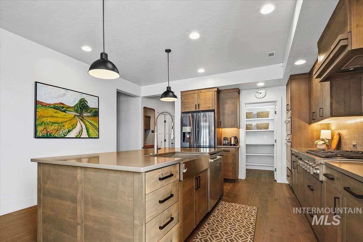 Kitchen featuring a kitchen island with sink, tasteful backsplash, stainless steel appliances, decorative light fixtures, and dark wood-type flooring