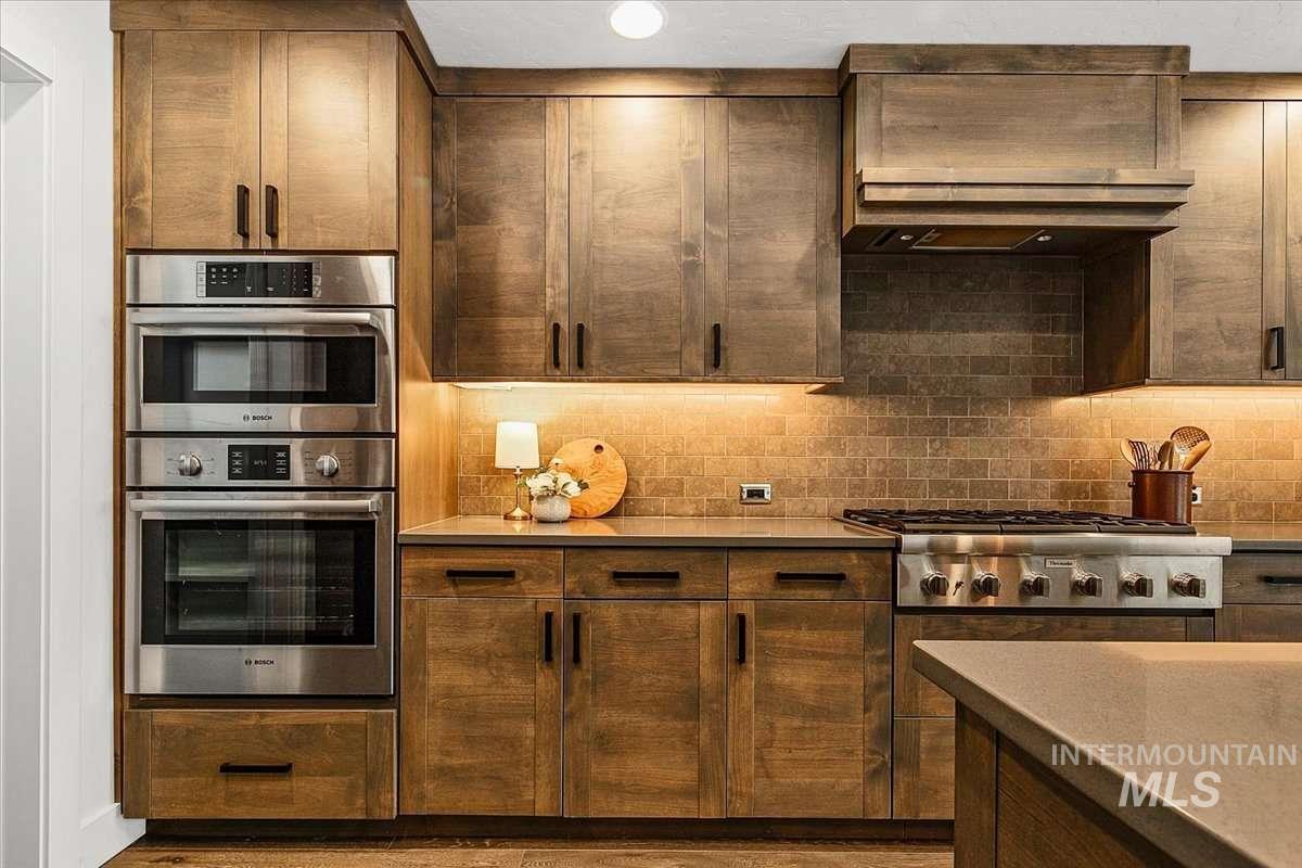 Kitchen with stainless steel appliances, exhaust hood, dark wood finish cabinets, decorative backsplash, and light stone counters