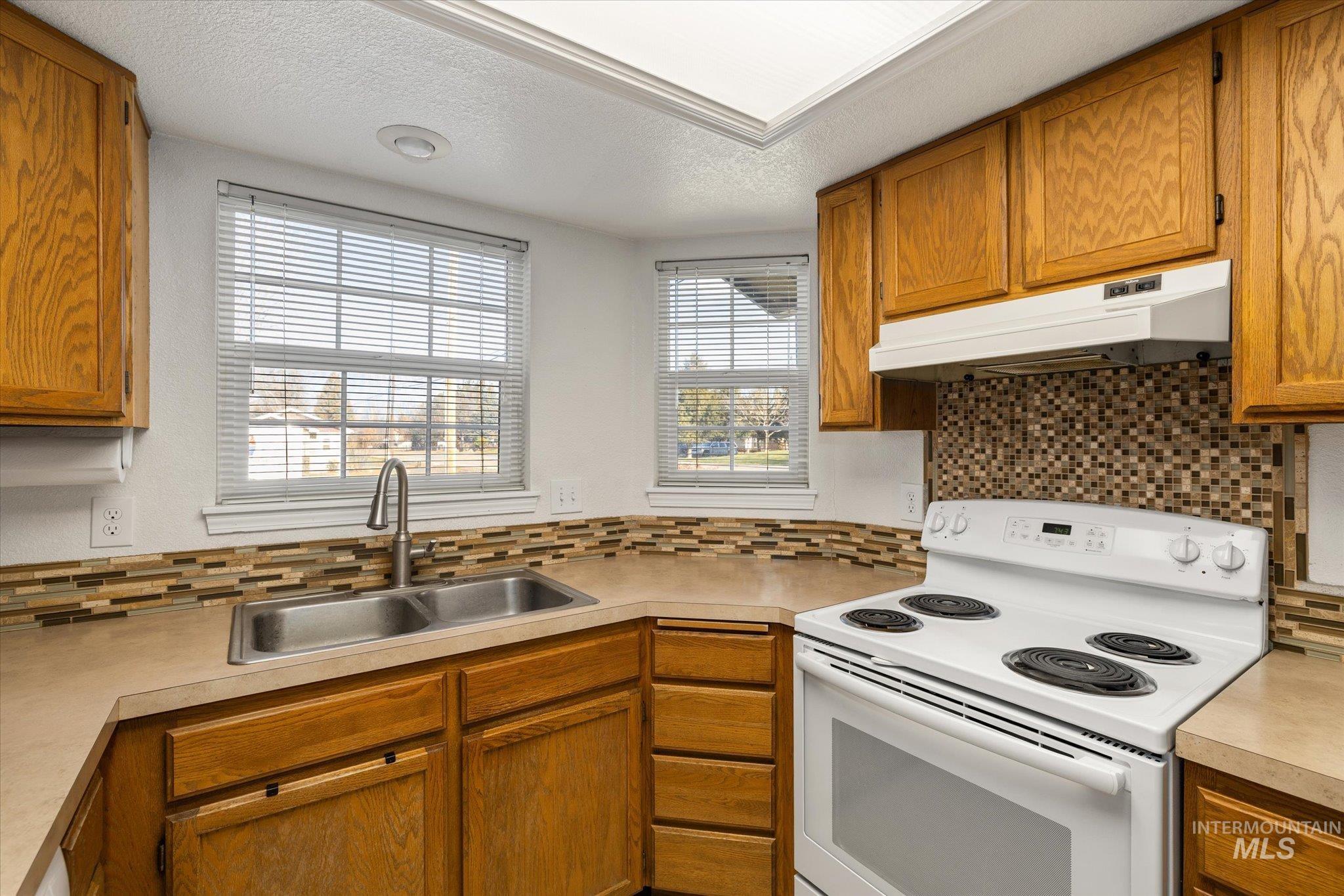Kitchen with white electric range, brown cabinetry, light countertops, under cabinet range hood, and a textured ceiling