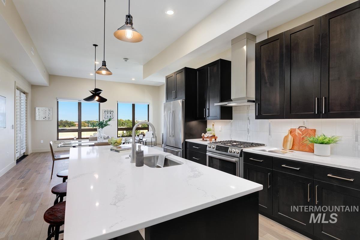 Kitchen with light wood-style flooring, decorative backsplash, an island with sink, appliances with stainless steel finishes, and wall chimney range hood