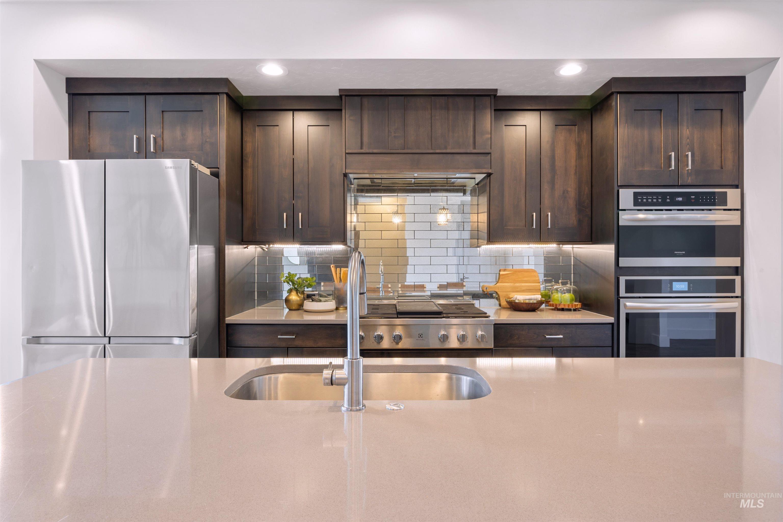 Kitchen featuring stainless steel appliances, dark wood finish cabinetry, light stone countertops, decorative backsplash, and recessed lighting