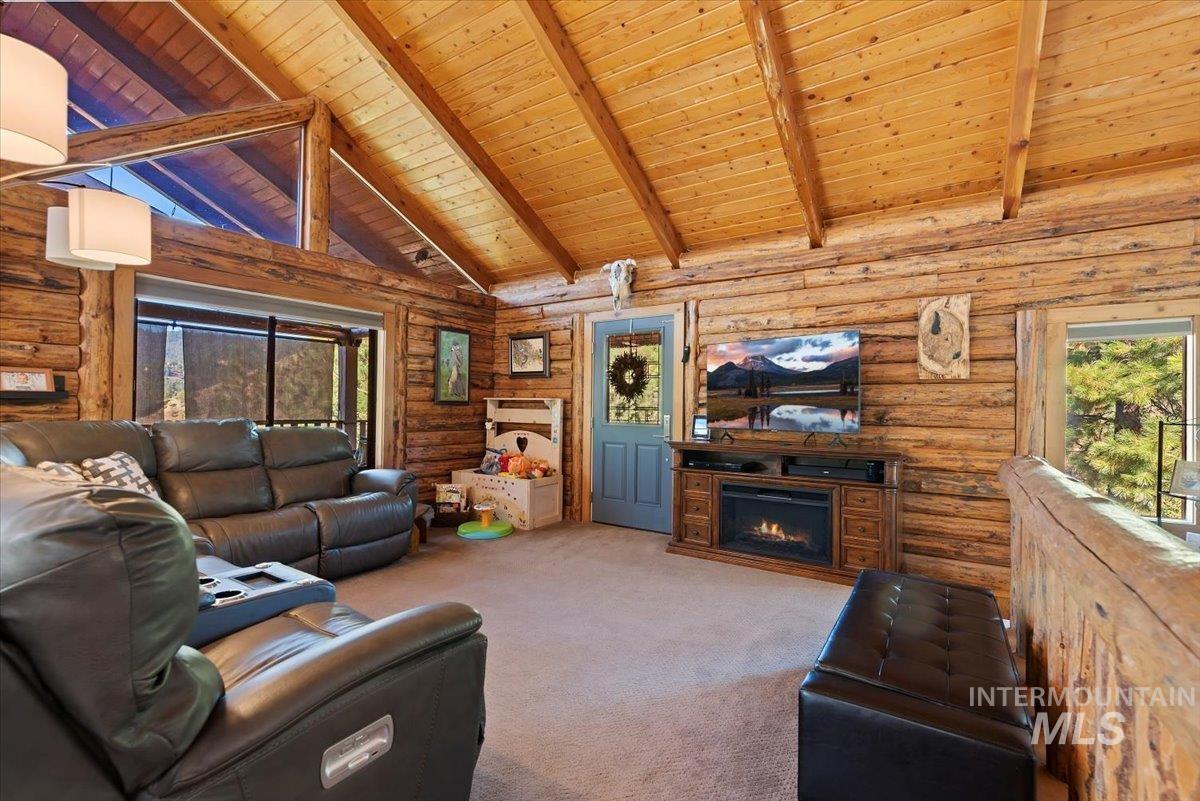 Living room featuring log walls, carpet flooring, a wood ceiling with exposed beams, a lit fireplace, and high vaulted ceiling