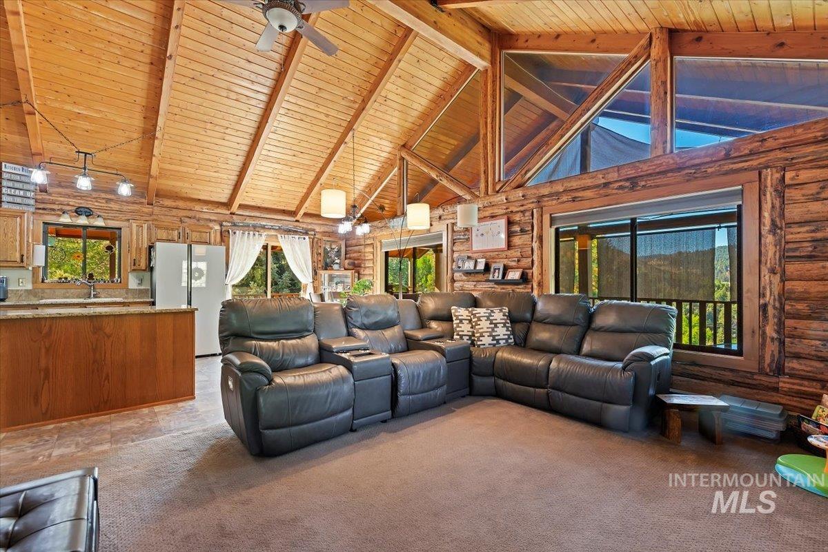 Living room with high vaulted ceiling, a ceiling fan, rustic walls, a wooden ceiling with exposed beams, and light colored carpet