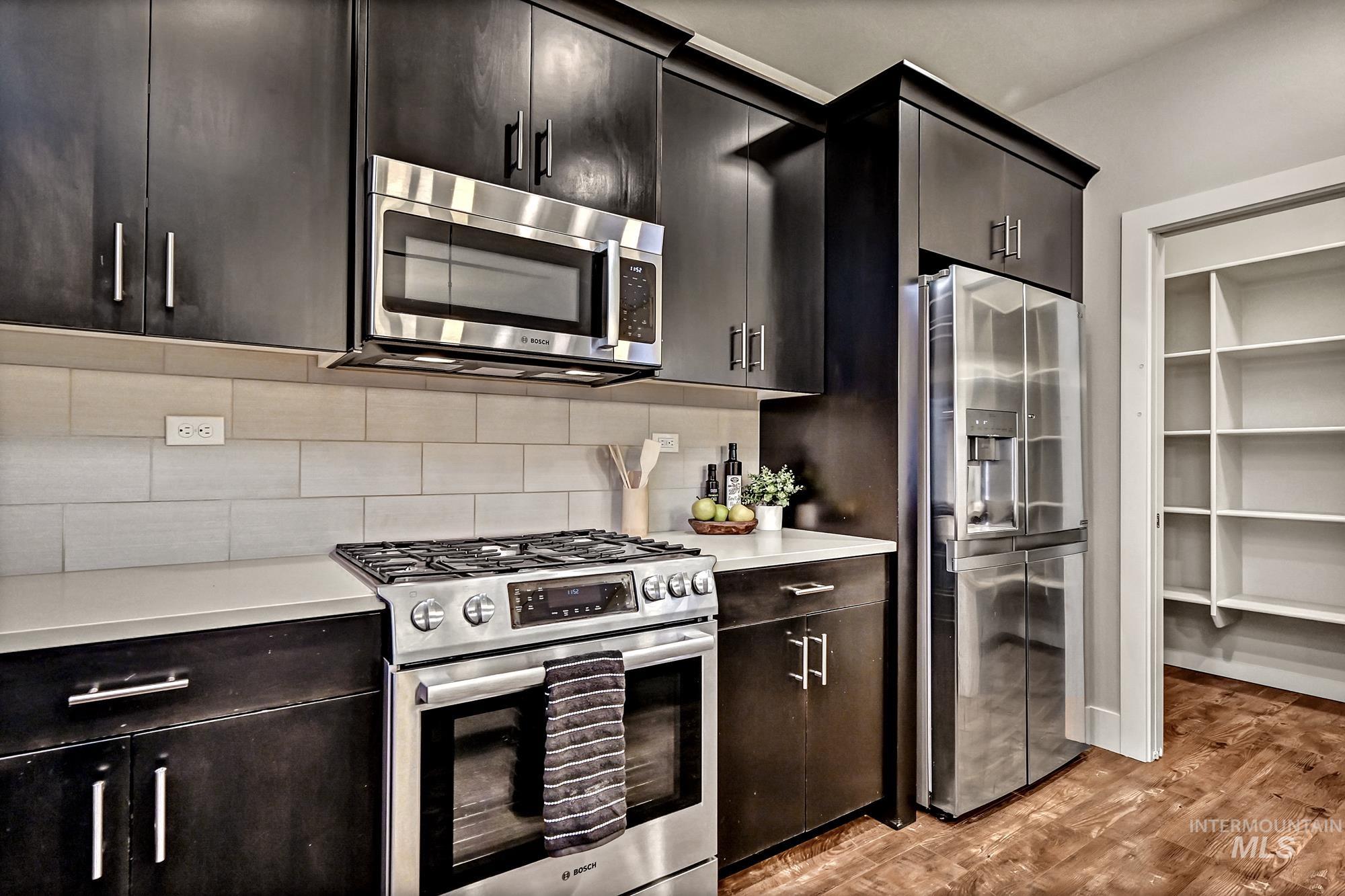 Kitchen featuring an island, appliances with stainless steel finishes, pendant light fixtures, tasteful tile backsplash, and dark wood-style flooring with the entry to a large pantry