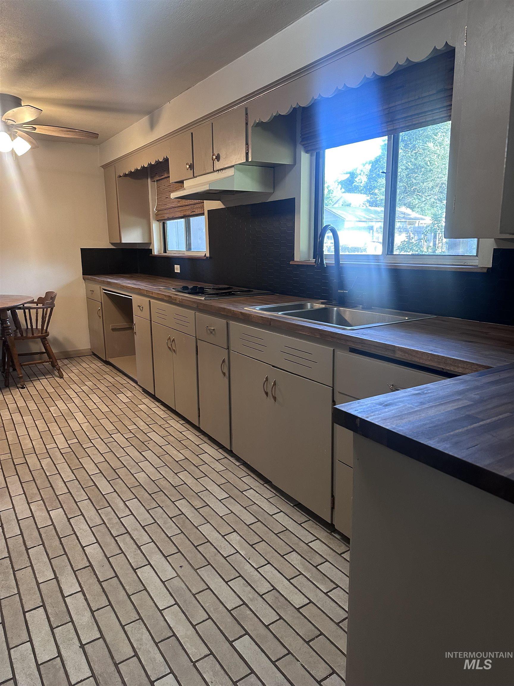 Kitchen featuring dark countertops, a ceiling fan, backsplash, under cabinet range hood, and brick patterned flooring