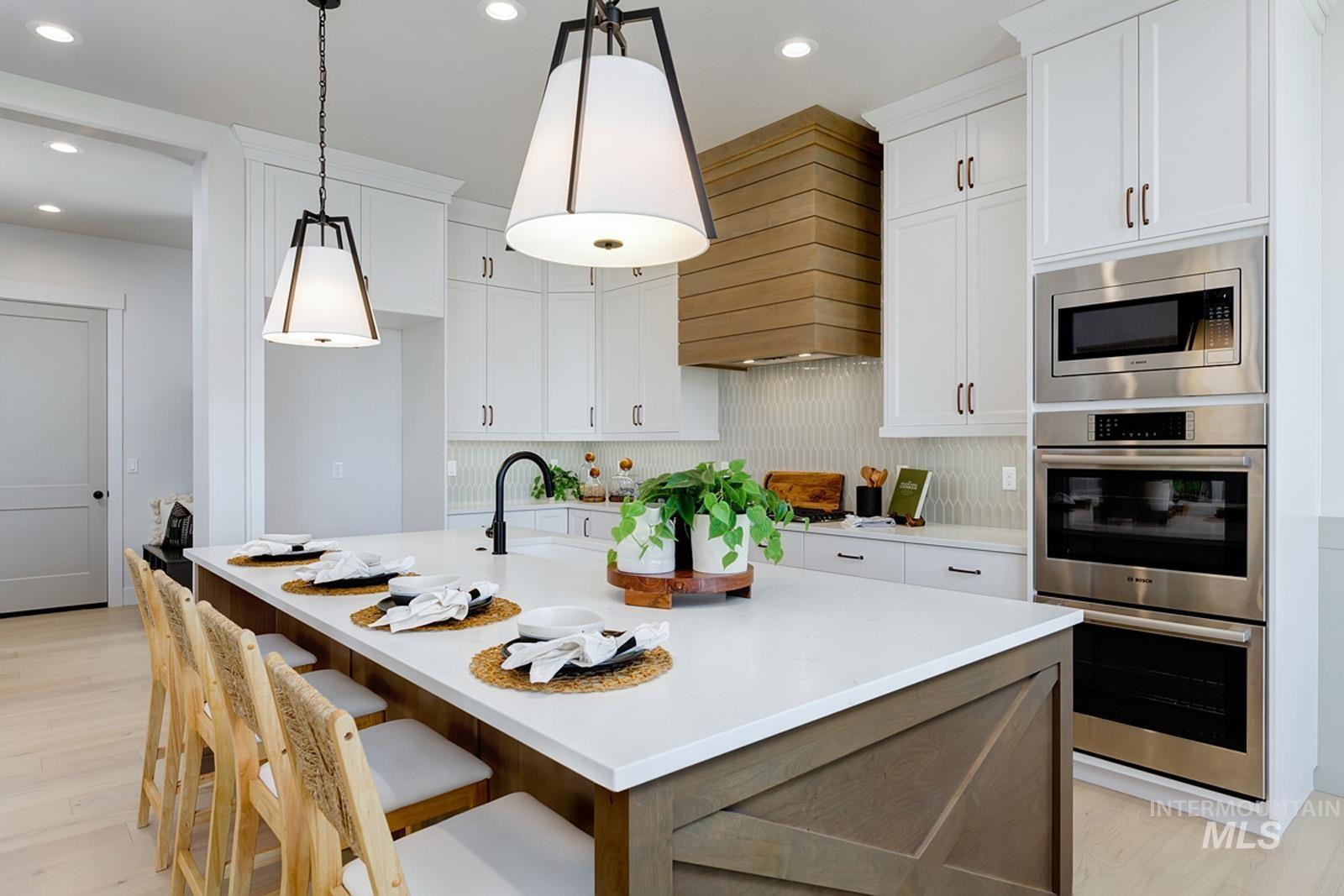 Kitchen featuring stainless steel appliances, a center island with sink, light wood-style floors, decorative light fixtures, and light stone counters