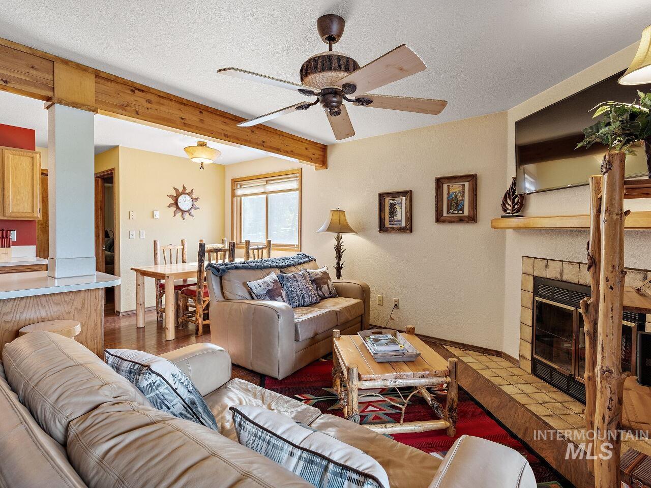 Living room featuring ceiling fan, beam ceiling, a tiled fireplace, wood finished floors, and a textured ceiling