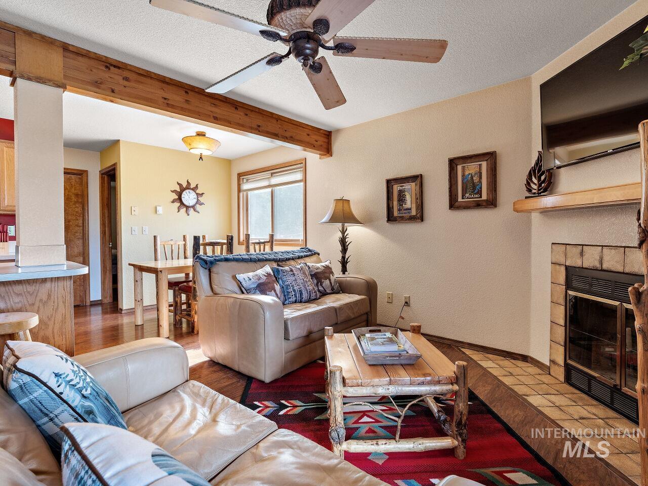 Living room featuring a ceiling fan, beam ceiling, a tile fireplace, a textured ceiling, and wood finished floors
