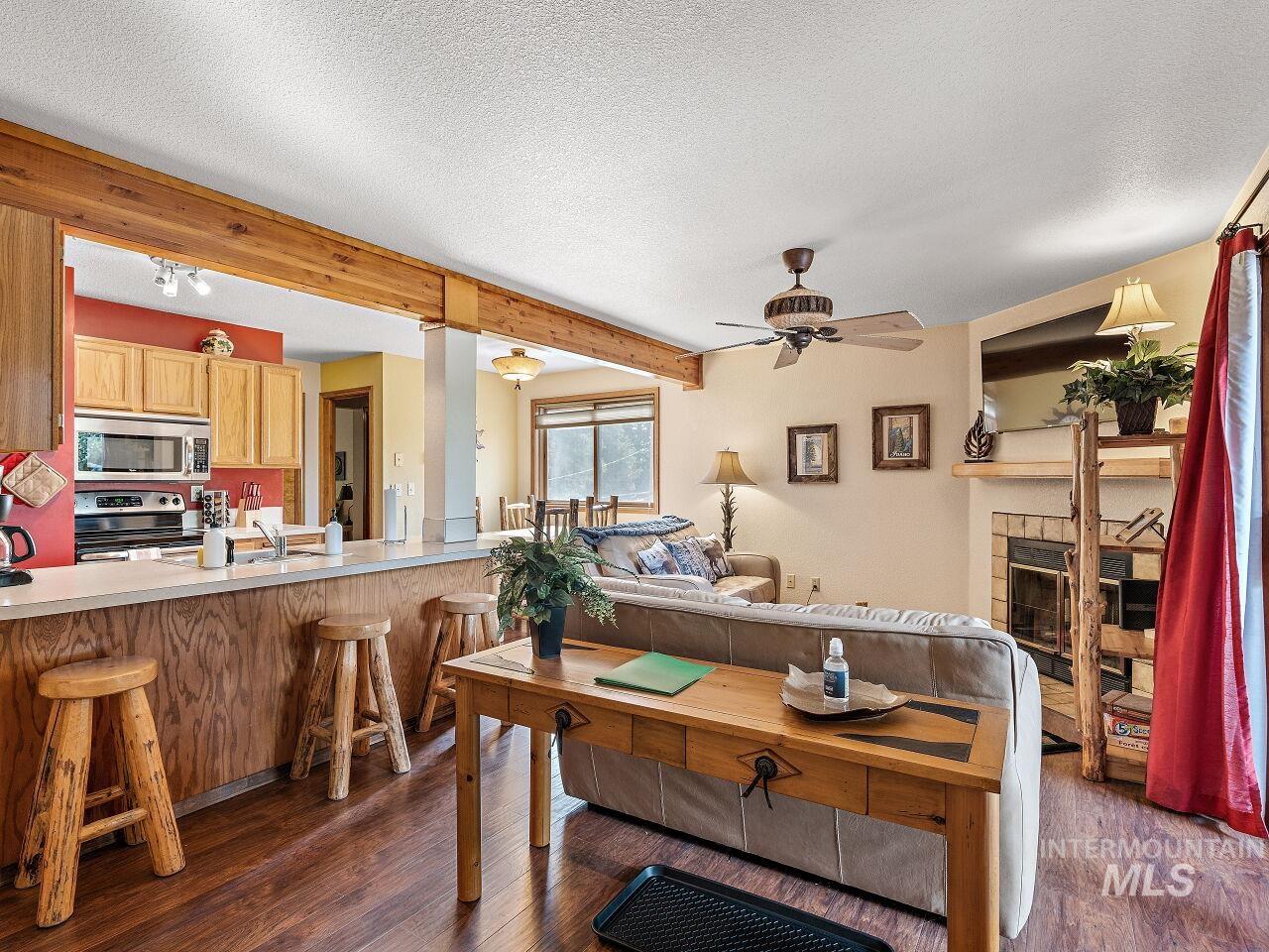 Living area featuring ceiling fan, dark wood-style floors, a tiled fireplace, beam ceiling, and a textured ceiling