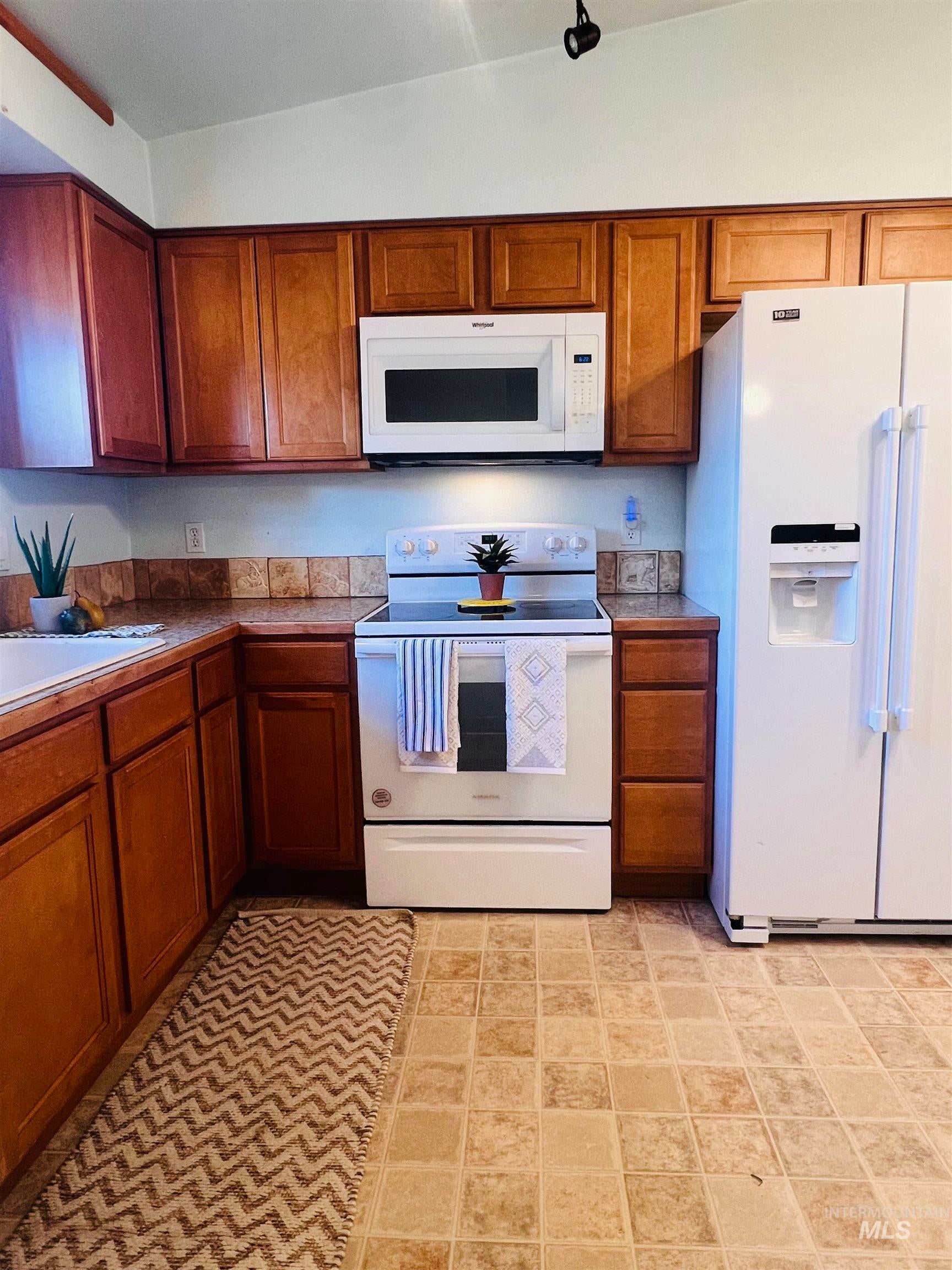 Kitchen featuring wood-finish cabinetry, white appliances, square tile backsplash, and light-toned square tile flooring