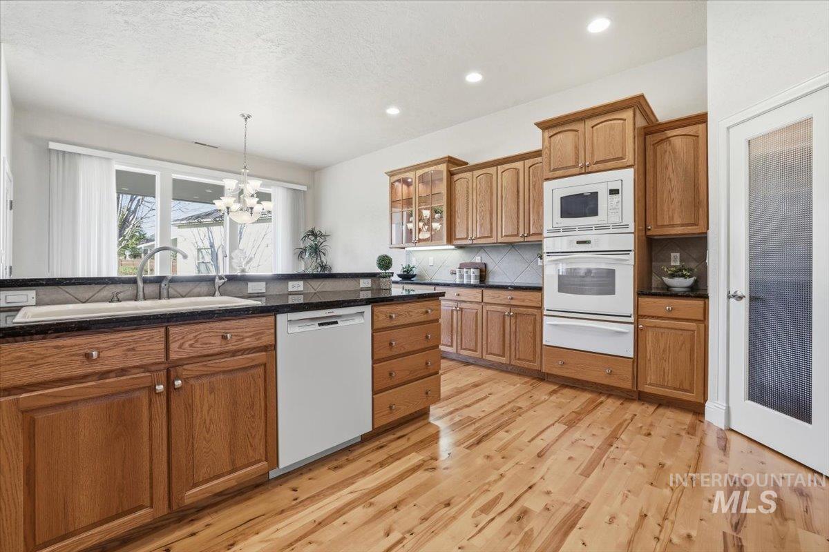 Kitchen with brown cabinets, glass insert cabinets, backsplash, white appliances, and a chandelier