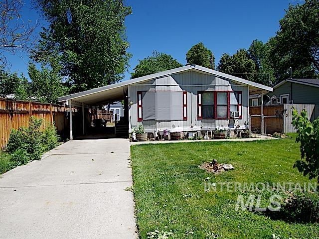Single-story residence featuring light gray siding with red trim accents, a covered carport, a concrete driveway, a front lawn, and wood fencing