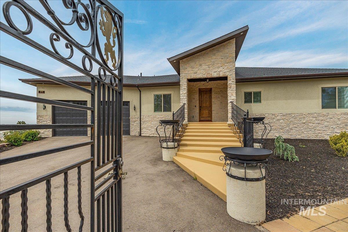Wrought iron entry gate opens to a paved driveway leading to a stone and stucco residence with a prominent wooden entry door and dual garage doors