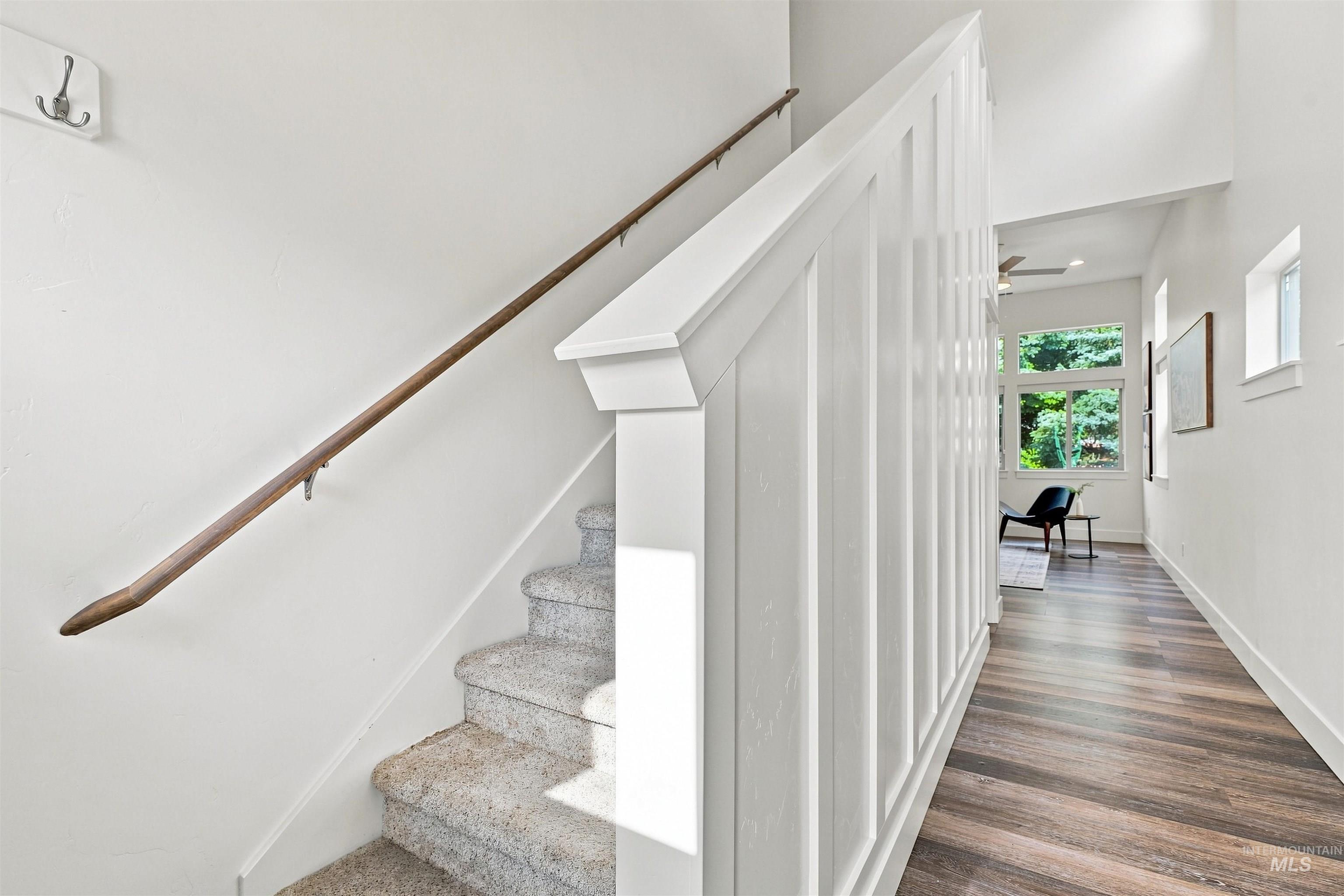 Carpeted staircase with a wood handrail, white board and batten wall paneling, and wood-finish flooring extending into a bright hallway with large windows