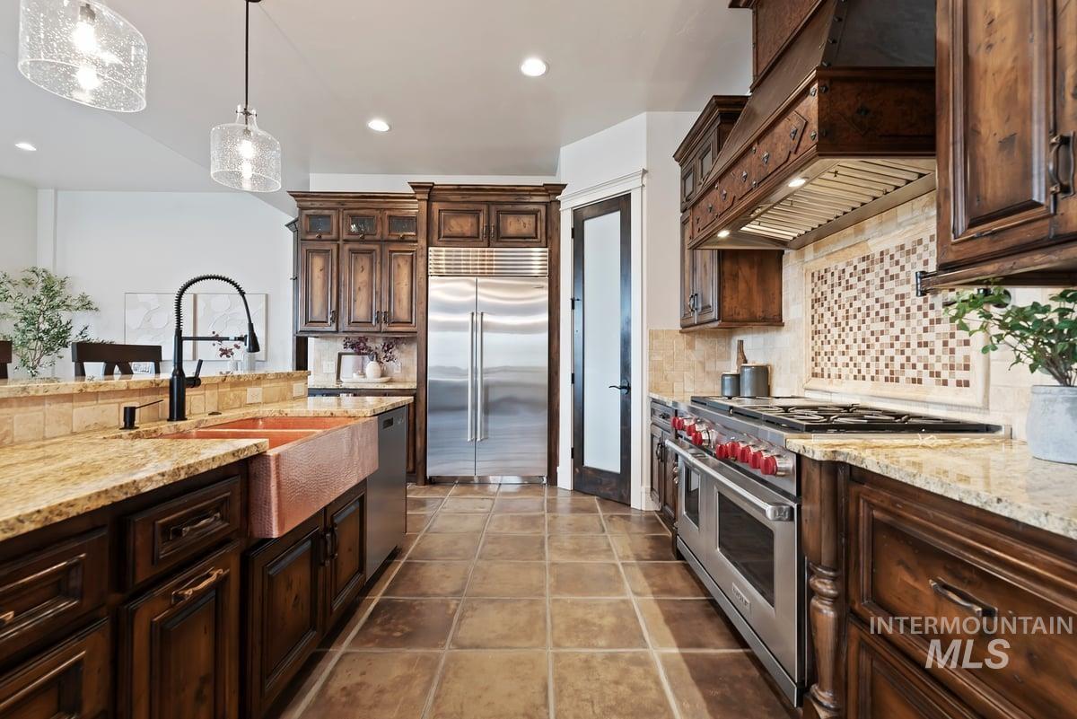 Kitchen featuring dark brown cabinets, light stone countertops, high quality appliances, custom range hood, and decorative light fixtures