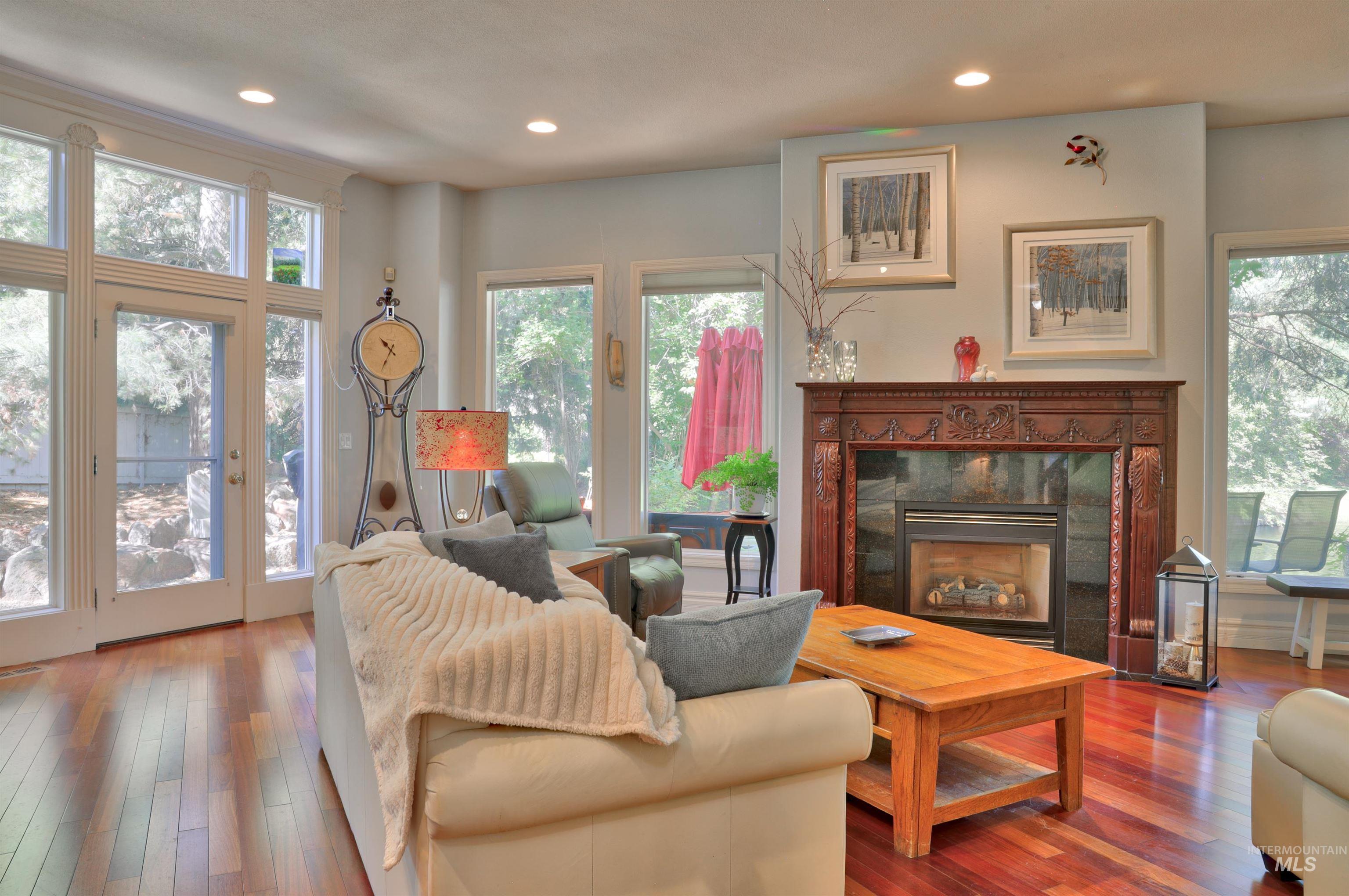 Spacious living area featuring polished wood-finish flooring, a detailed fireplace with an ornate wood mantel, multiple large windows, and recessed lighting