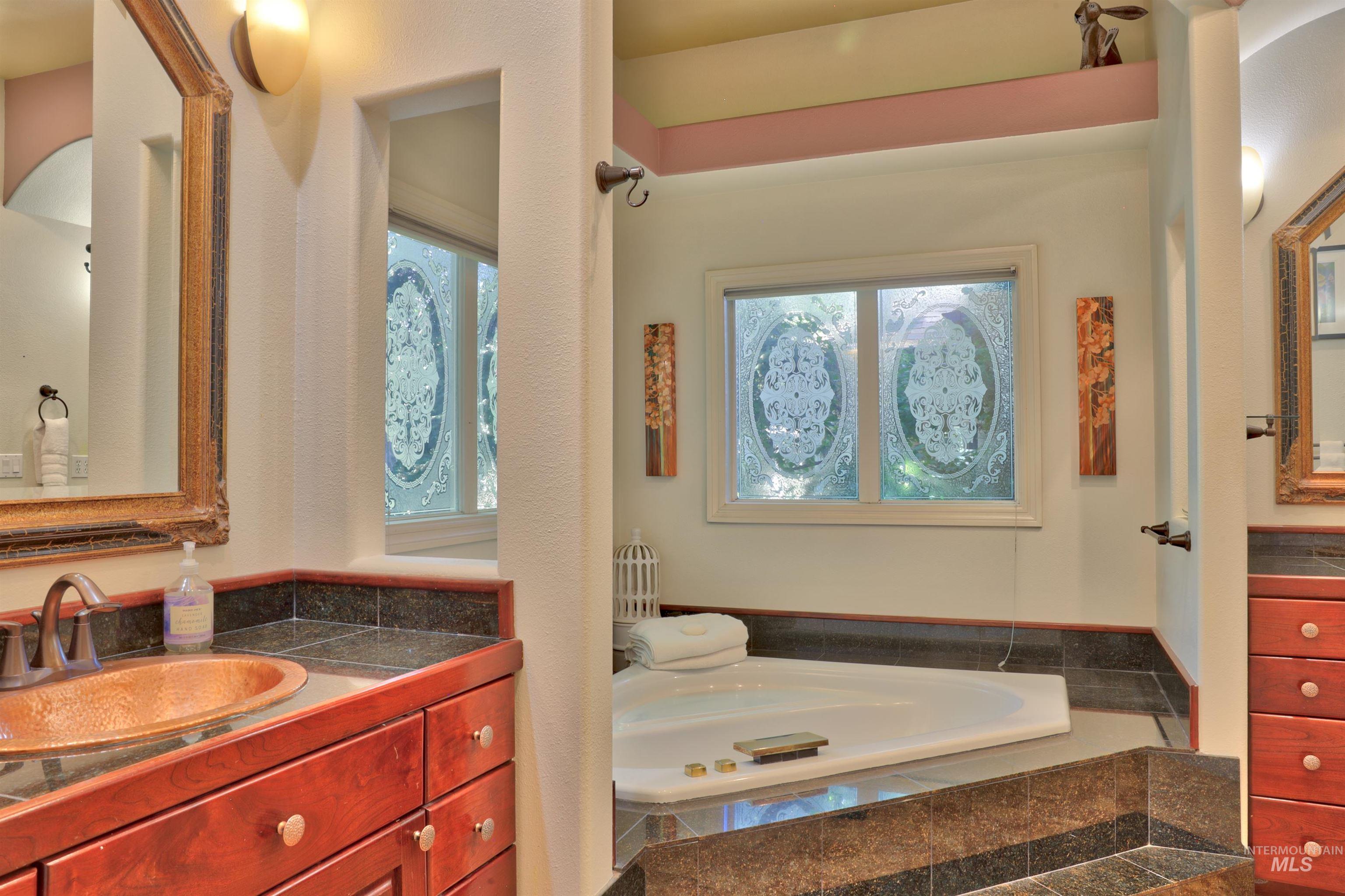 Bathroom featuring a built-in corner soaking tub with dark stone surround, custom wood cabinetry with a copper vessel sink, granite countertops, and decorative privacy windows