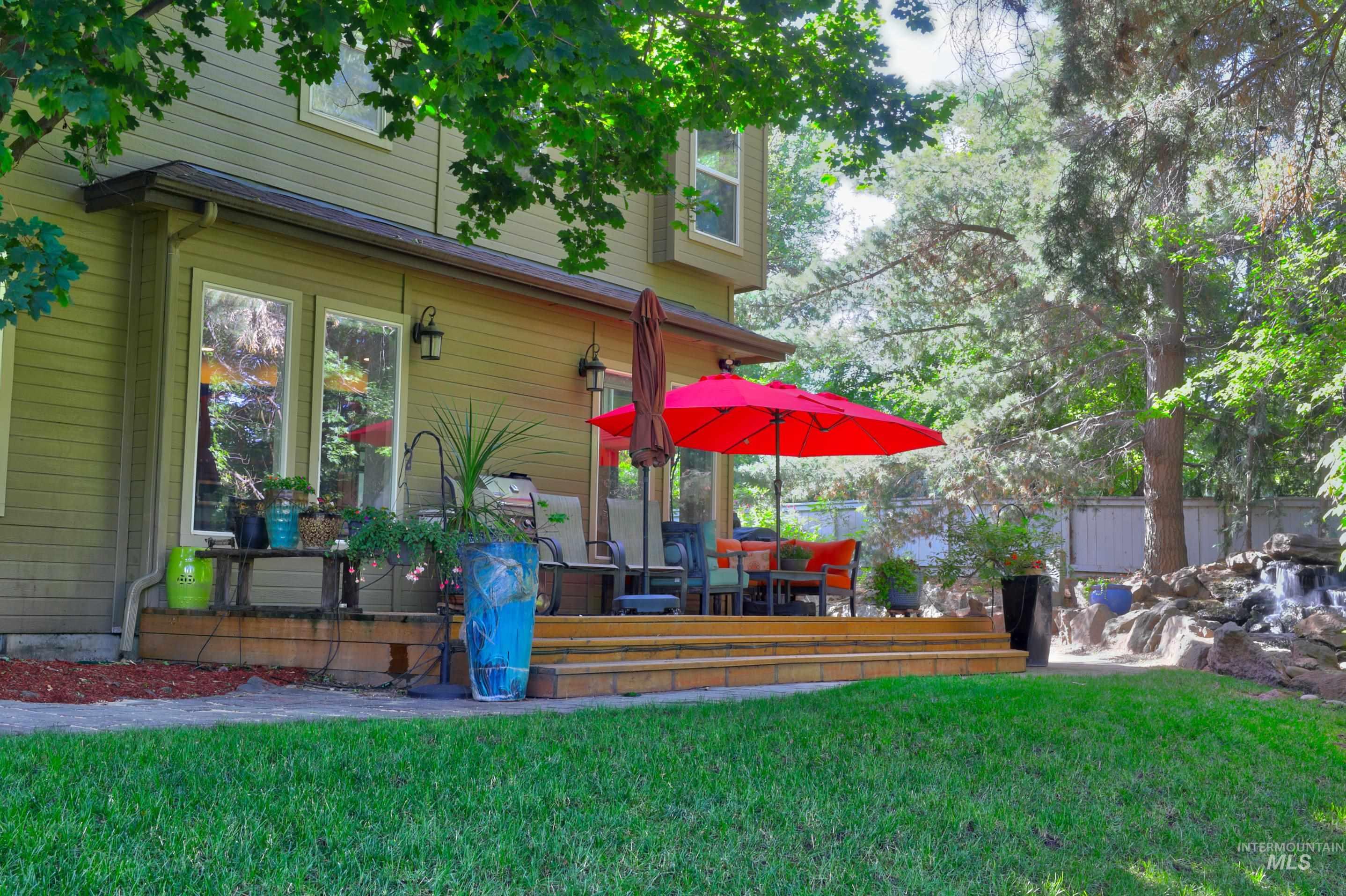 Expansive wooden deck with direct access to the interior, featuring a tiered rock water feature, mature trees, and a manicured lawn