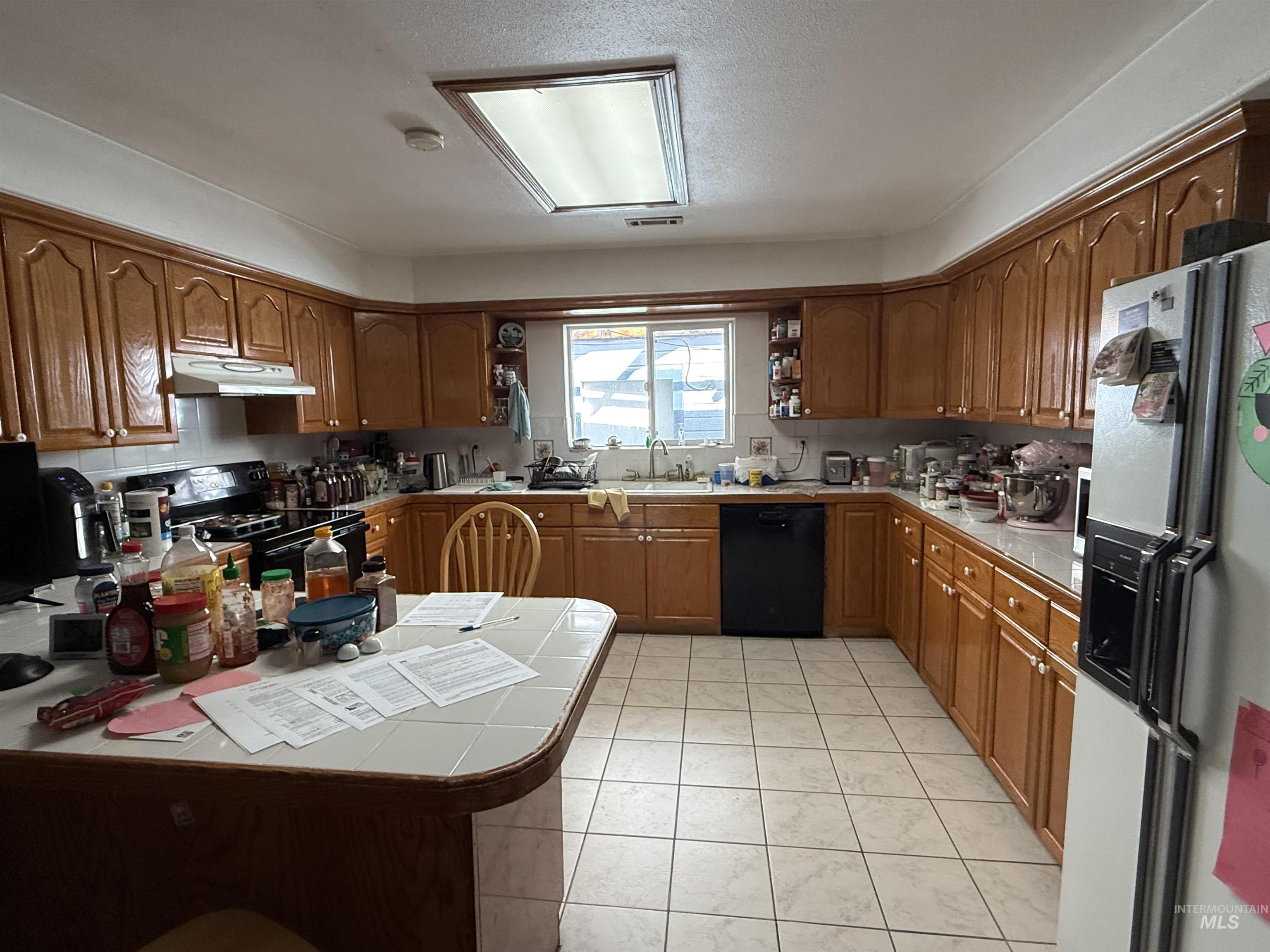 Kitchen featuring tile countertops, brown cabinets, black appliances, light tile patterned floors, and open shelves