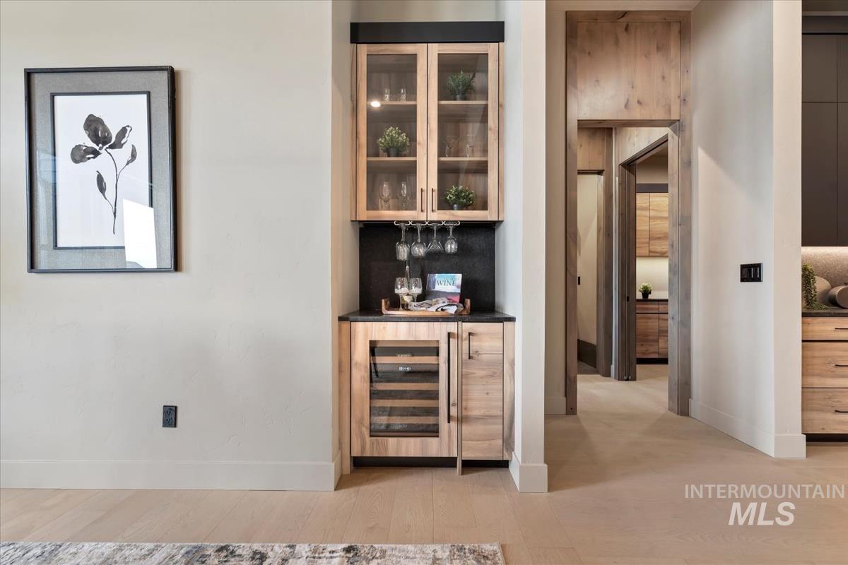 Indoor dry bar with glass insert cabinets, light wood-type flooring, wine cooler, light brown cabinets, and dark stone countertops