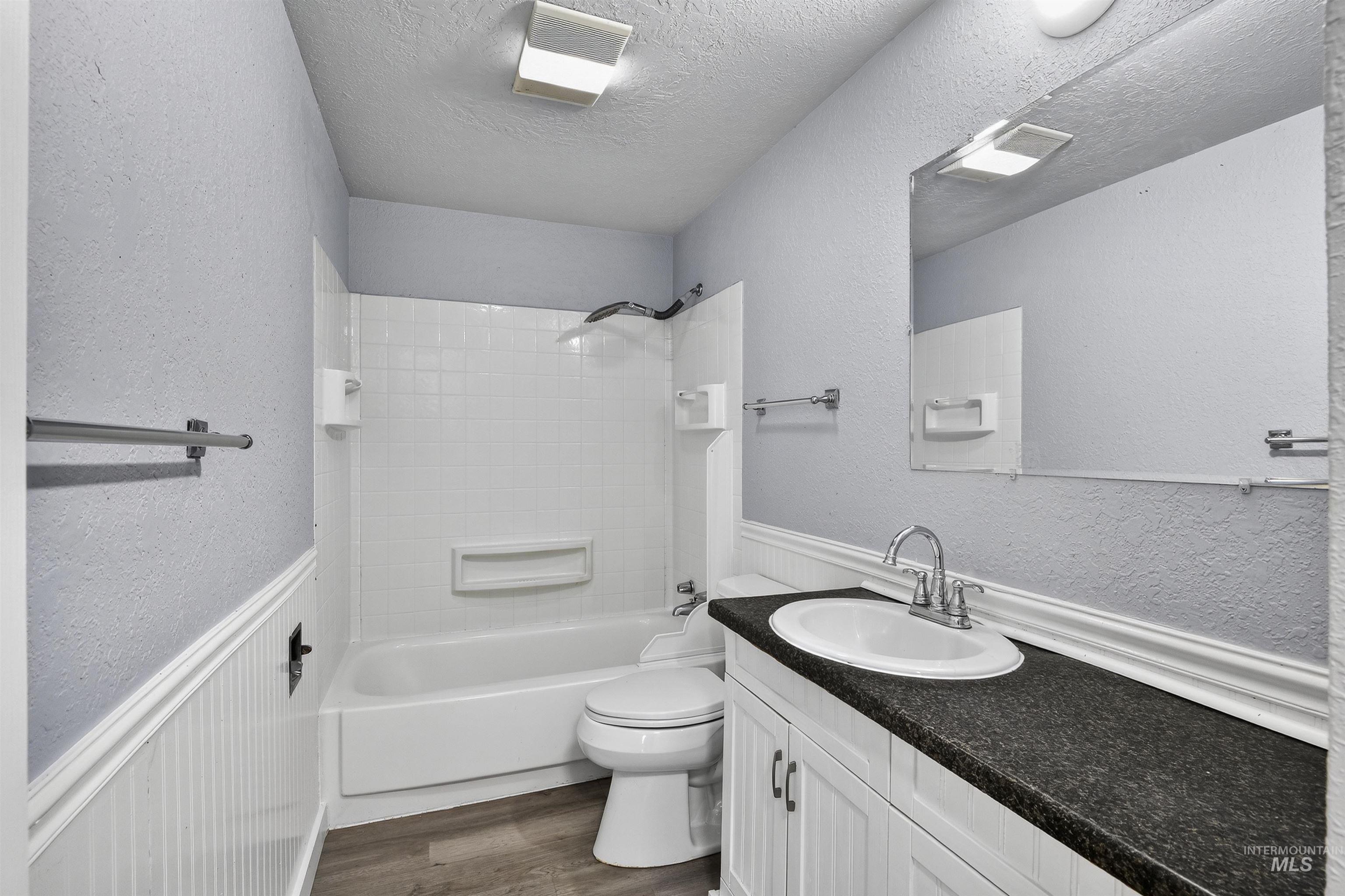 Full bath with a textured wall, wainscoting, shower / bathtub combination, vanity, and dark wood-style floors