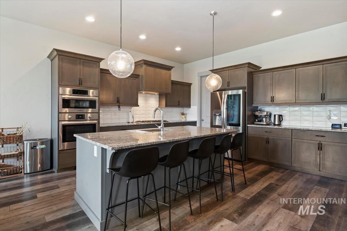 Kitchen featuring a large island with seating, a stainless steel appliance suite including double ovens and a built-in microwave, wood-finish flooring, full-height cabinetry with dark hardware, and a textured white tile backsplash
