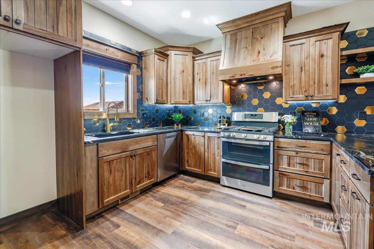 Kitchen featuring appliances with stainless steel finishes, light wood-style floors, custom exhaust hood, backsplash, and dark stone counters