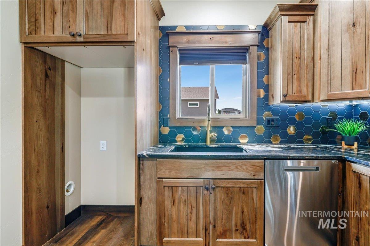 Kitchen featuring stainless steel dishwasher, backsplash, dark wood-style floors, dark stone counters, and brown cabinets