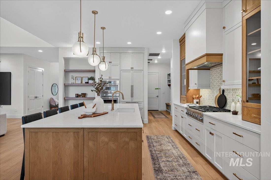 Kitchen featuring decorative light fixtures, light stone counters, a kitchen island with sink, backsplash, and light wood-style floors