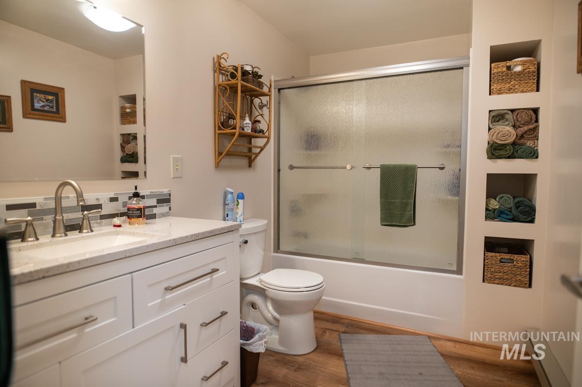 Full bath featuring vanity, combined bath / shower with glass door, wood finished floors, baseboard heating, and decorative backsplash