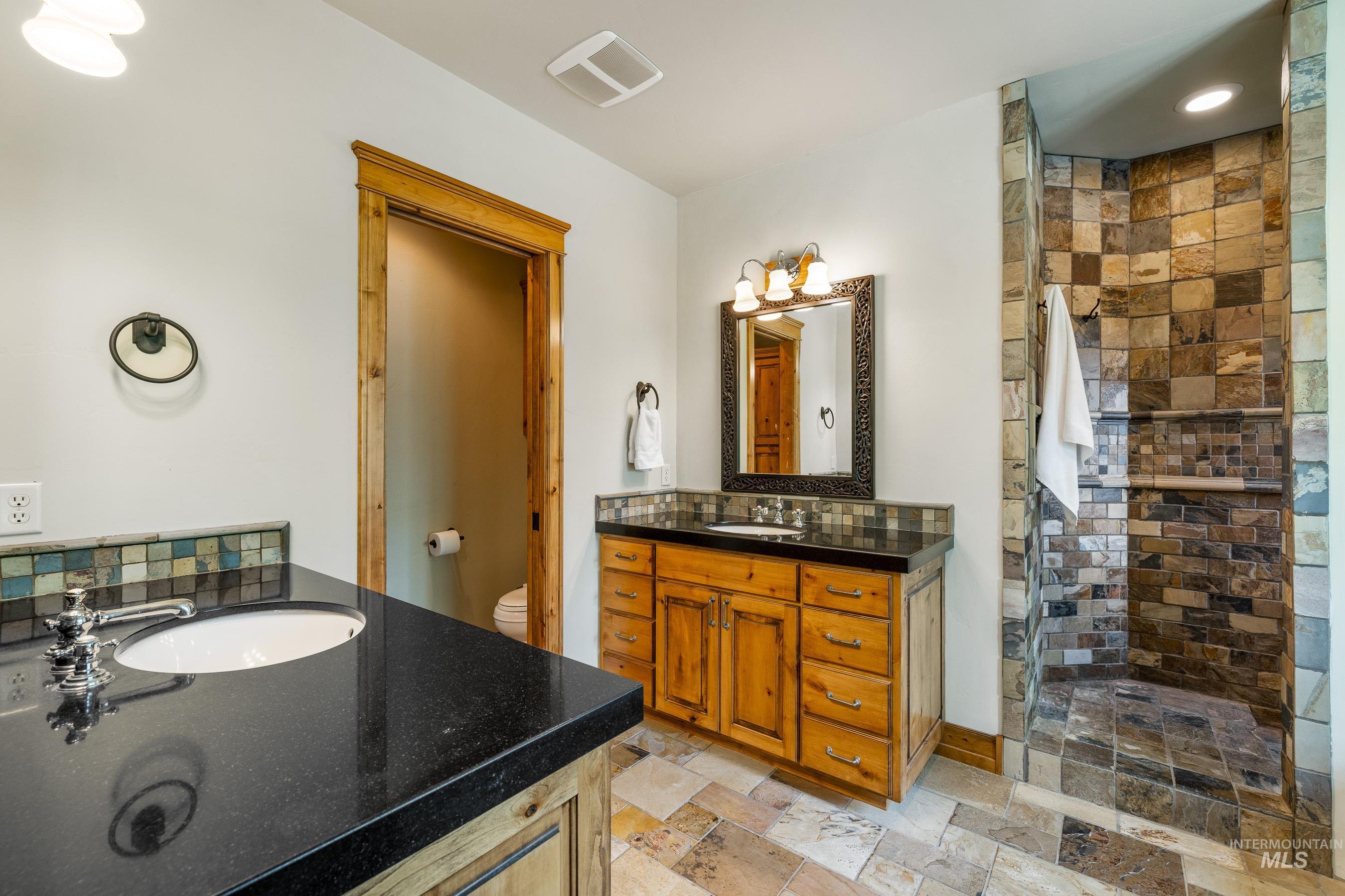 Bathroom featuring two vanities, a stall shower, backsplash, stone tile floors, and recessed lighting