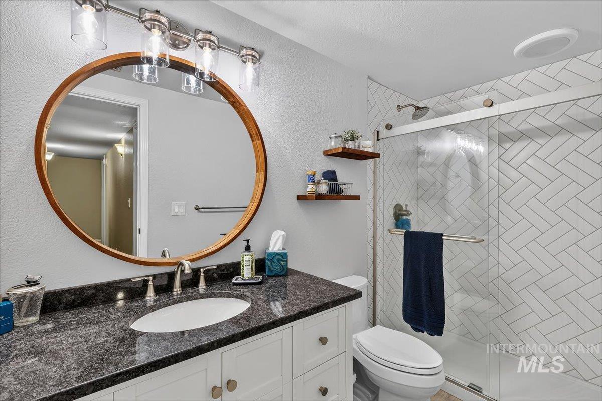 Bathroom featuring a dark speckled countertop vanity with an undermount sink, a circular wood-framed mirror, contemporary overhead lighting, and a walk-in shower with herringbone-patterned tile and a glass enclosure