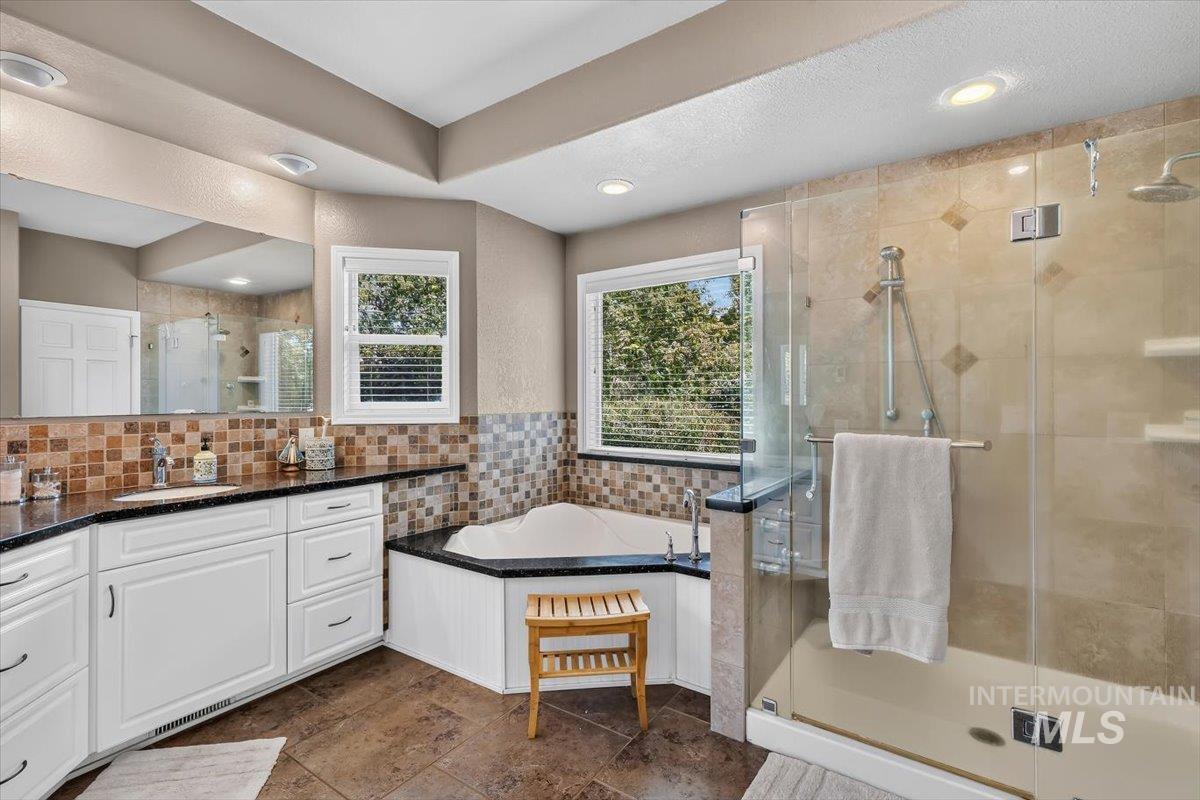Spacious bathroom featuring a corner soaking tub, glass-enclosed shower with multiple showerheads, dual-sink vanity with white cabinetry and dark countertops, and tile flooring