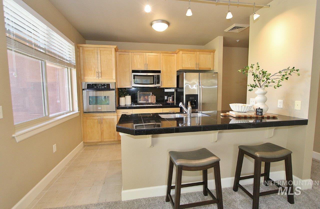 Kitchen with light brown cabinetry, a peninsula, appliances with stainless steel finishes, a breakfast bar, and tile counters