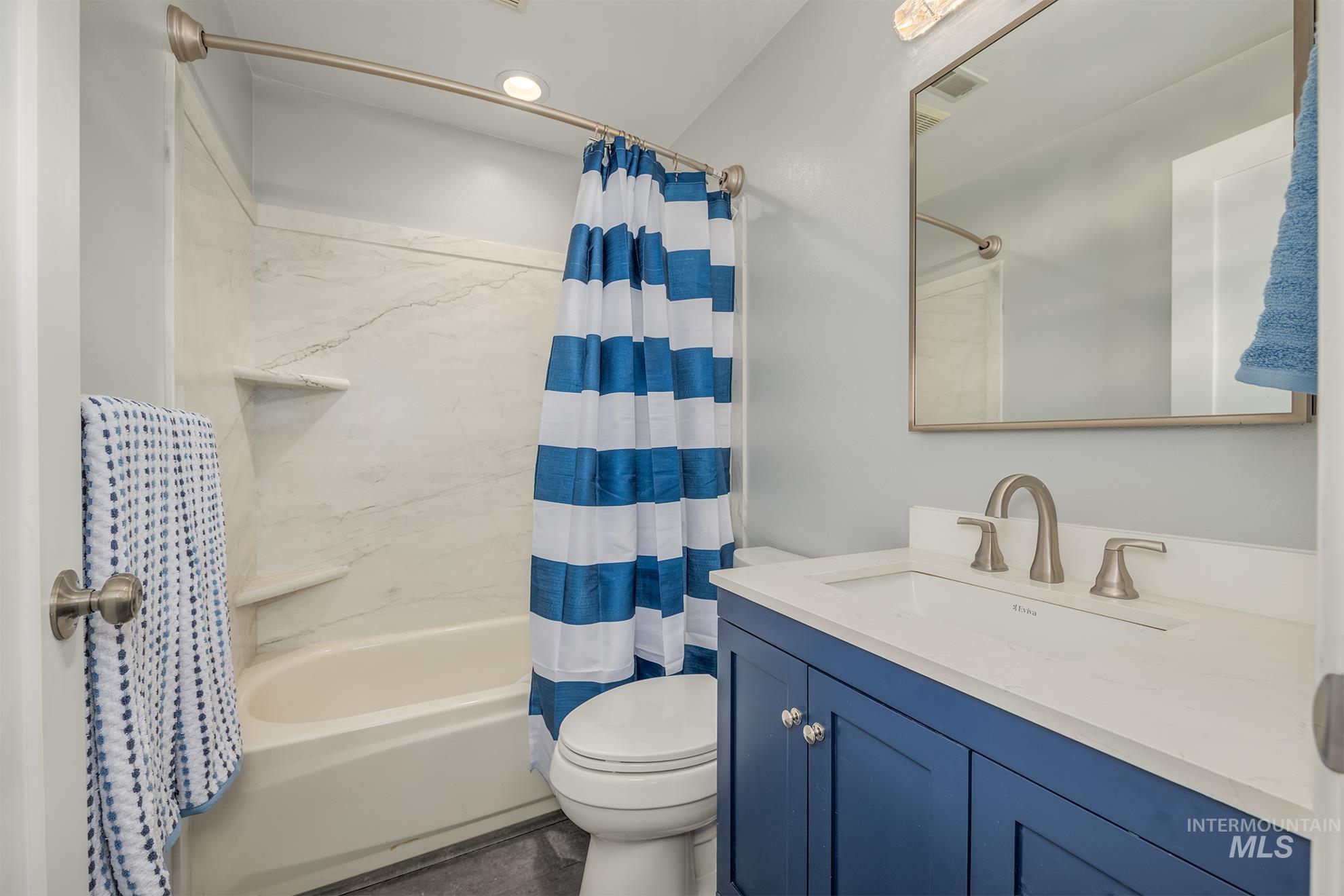 Bathroom featuring a white tub with a white-veined surround, a toilet, a blue vanity with a white countertop and integrated sink, a brushed nickel faucet, and a rectangular mirror with a brushed nickel frame