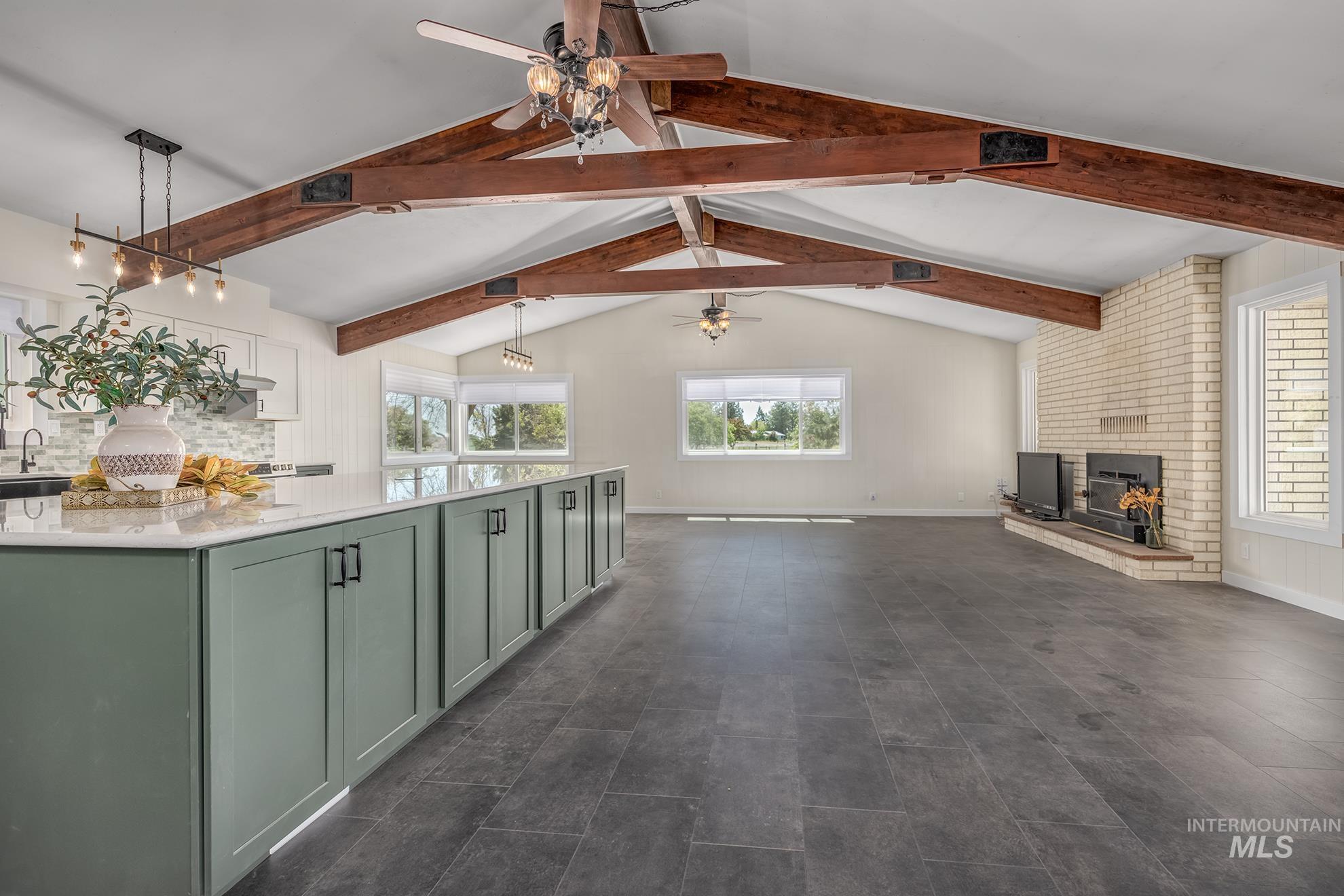 Expansive open-concept room featuring vaulted ceilings with exposed wood beams, a prominent kitchen island with sage green cabinetry and a light-toned countertop, and dark-toned plank flooring