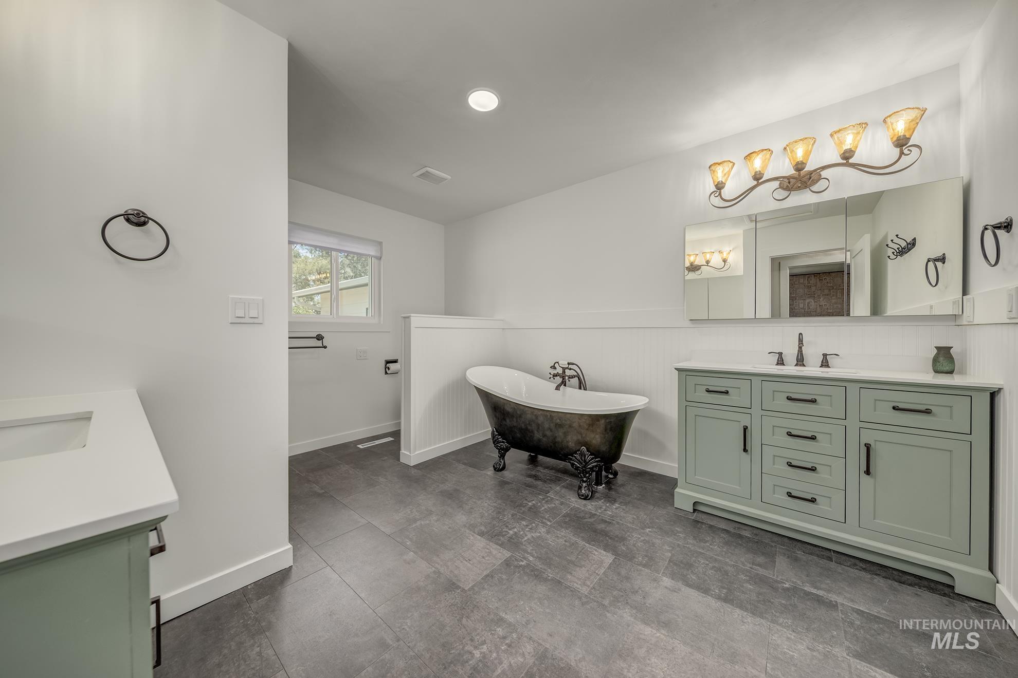 Spacious bathroom featuring a freestanding clawfoot tub, dual sage green vanities with integrated sinks and white countertops, dark floor tiling, and a large mirrored medicine cabinet with decorative lighting