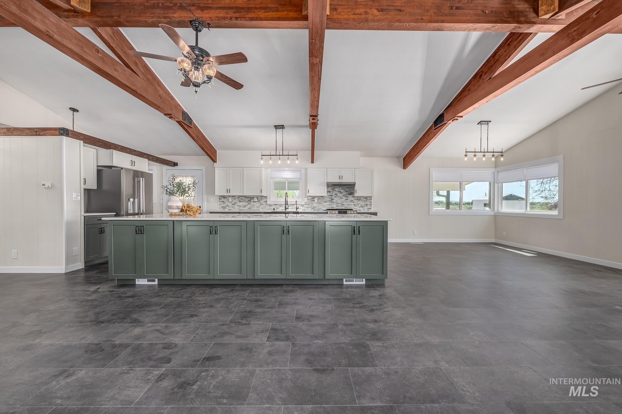 Spacious kitchen and living area featuring vaulted ceilings with exposed wood beams, a large kitchen island with sage green cabinetry and a white countertop, stainless steel appliances, and dark tile flooring