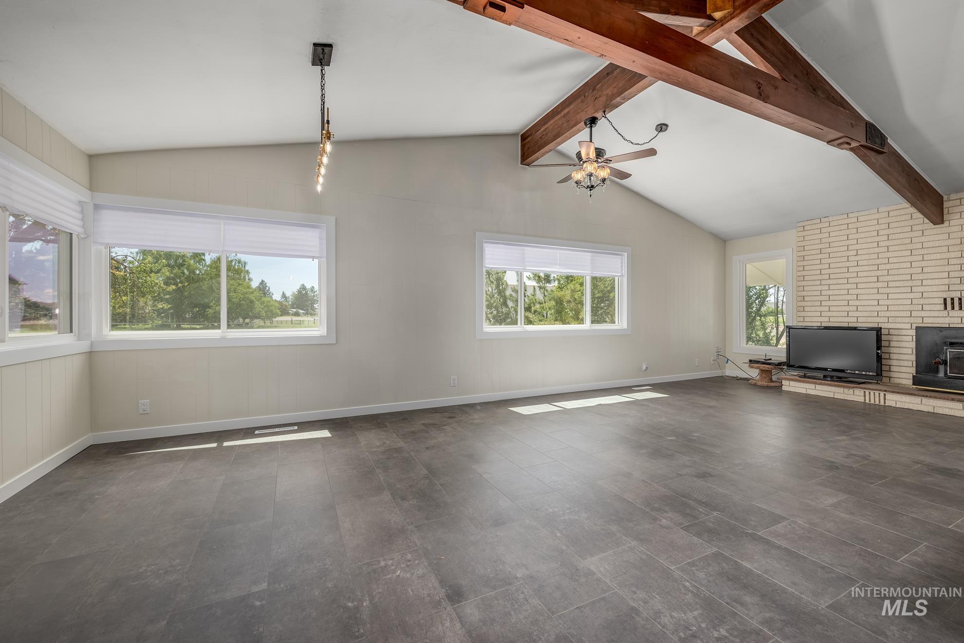 Spacious room featuring vaulted ceilings with exposed wood beams, wood-finish flooring, a brick fireplace with hearth, and multiple windows