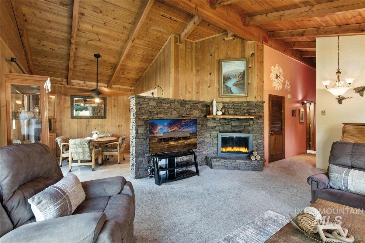 Living room with wooden walls, a wood ceiling with exposed beams, a stone fireplace, high vaulted ceiling, and carpet floors