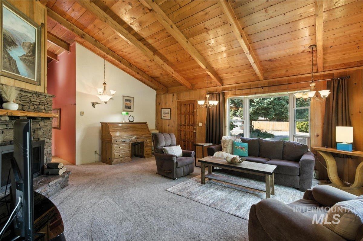 Carpeted living area featuring a chandelier, a stone fireplace, a wood ceiling with exposed beams, wood walls, and high vaulted ceiling