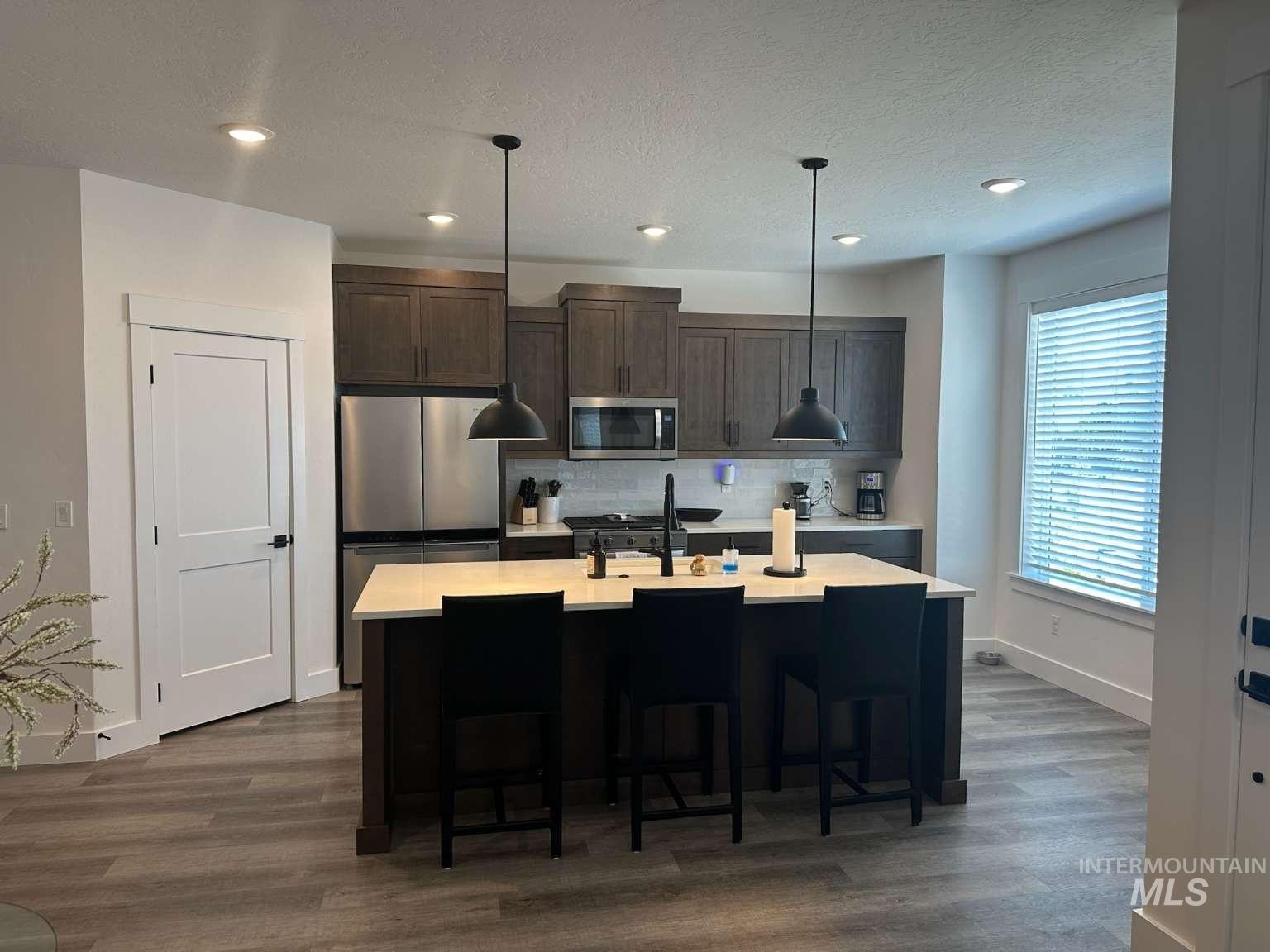 Kitchen with dark brown cabinetry, stainless steel appliances, a center island with sink, decorative light fixtures, and dark wood-style floors
