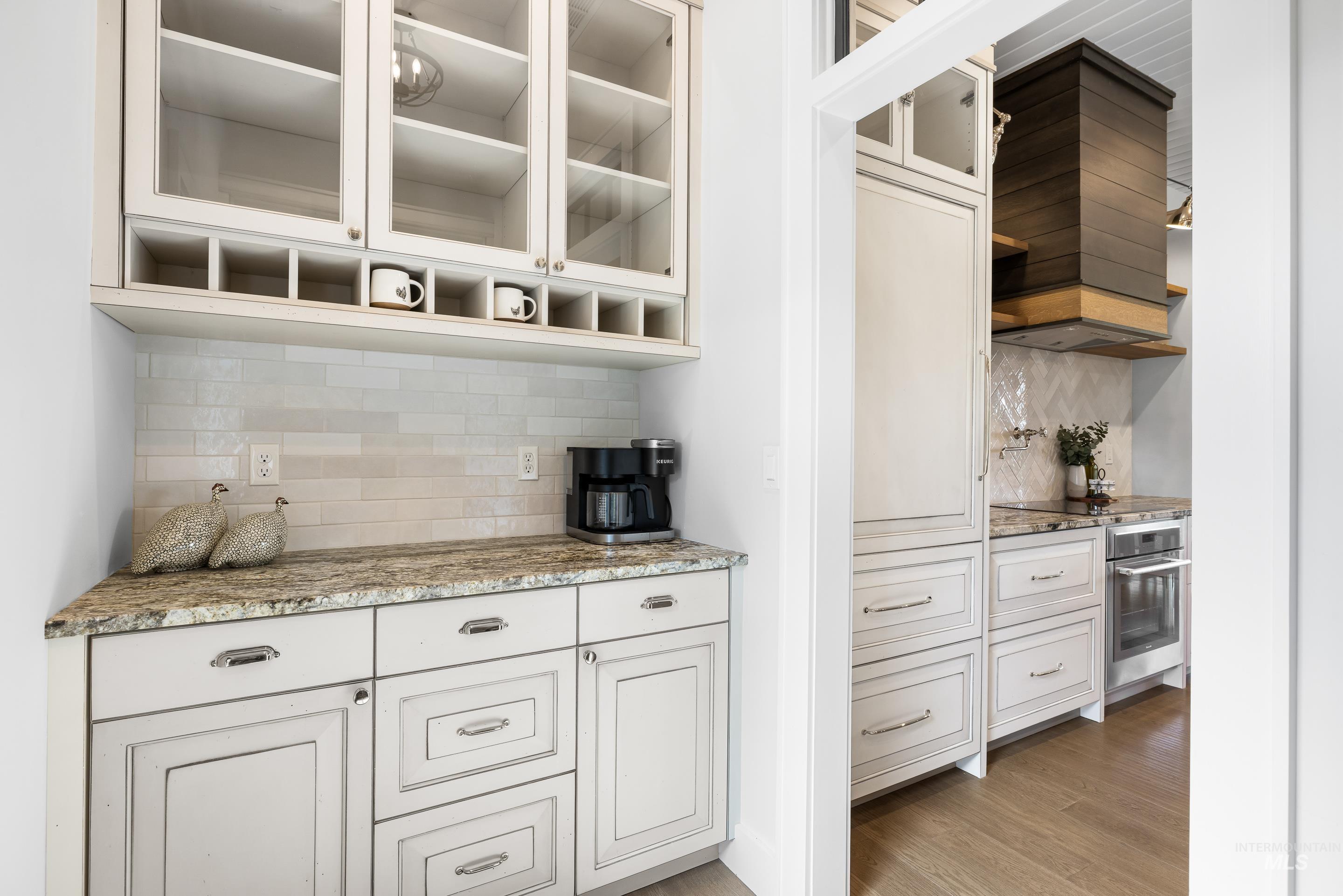 Bar area featuring decorative backsplash, light stone counters, glass insert cabinets, light wood-style flooring, and custom range hood