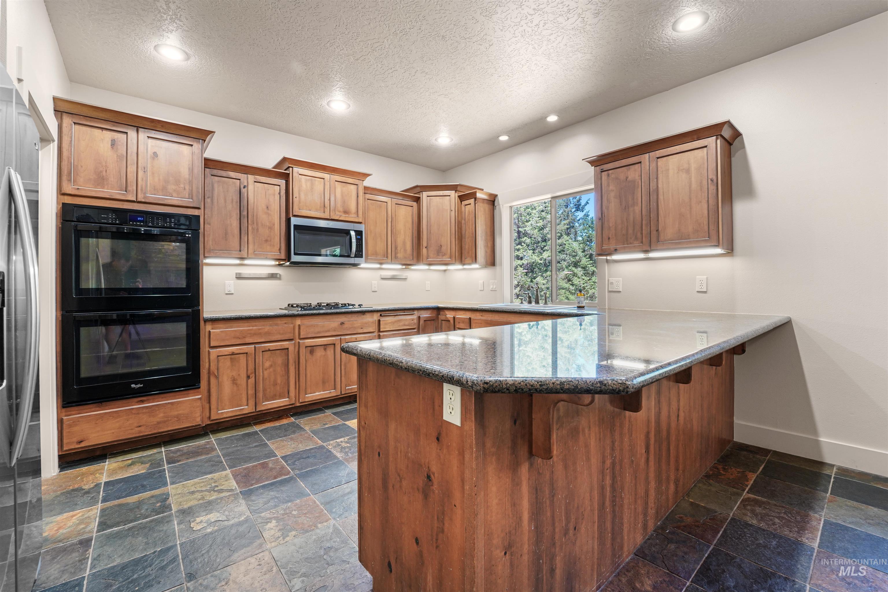 Kitchen with stainless steel microwave, a textured ceiling, stone tile floors, gas cooktop, and brown cabinets