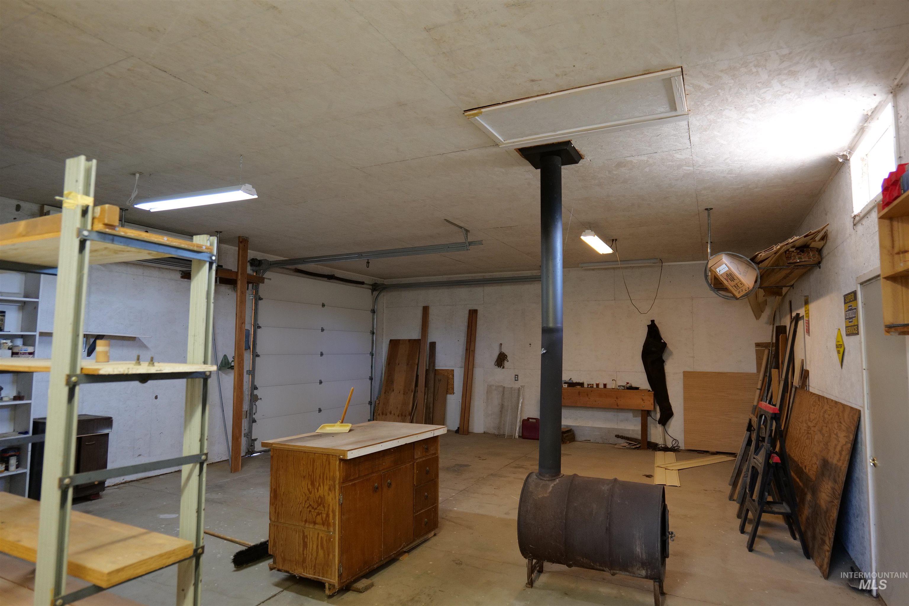 Spacious shop interior featuring concrete flooring, a large overhead door, fluorescent lighting, and a drum stove with an exhaust pipe