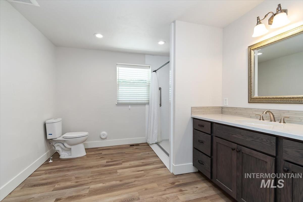 Bathroom featuring wood-finish flooring, a white vanity with an integrated sink and brushed nickel faucet, an ornate framed mirror, a glass-enclosed shower, and a window with horizontal blinds