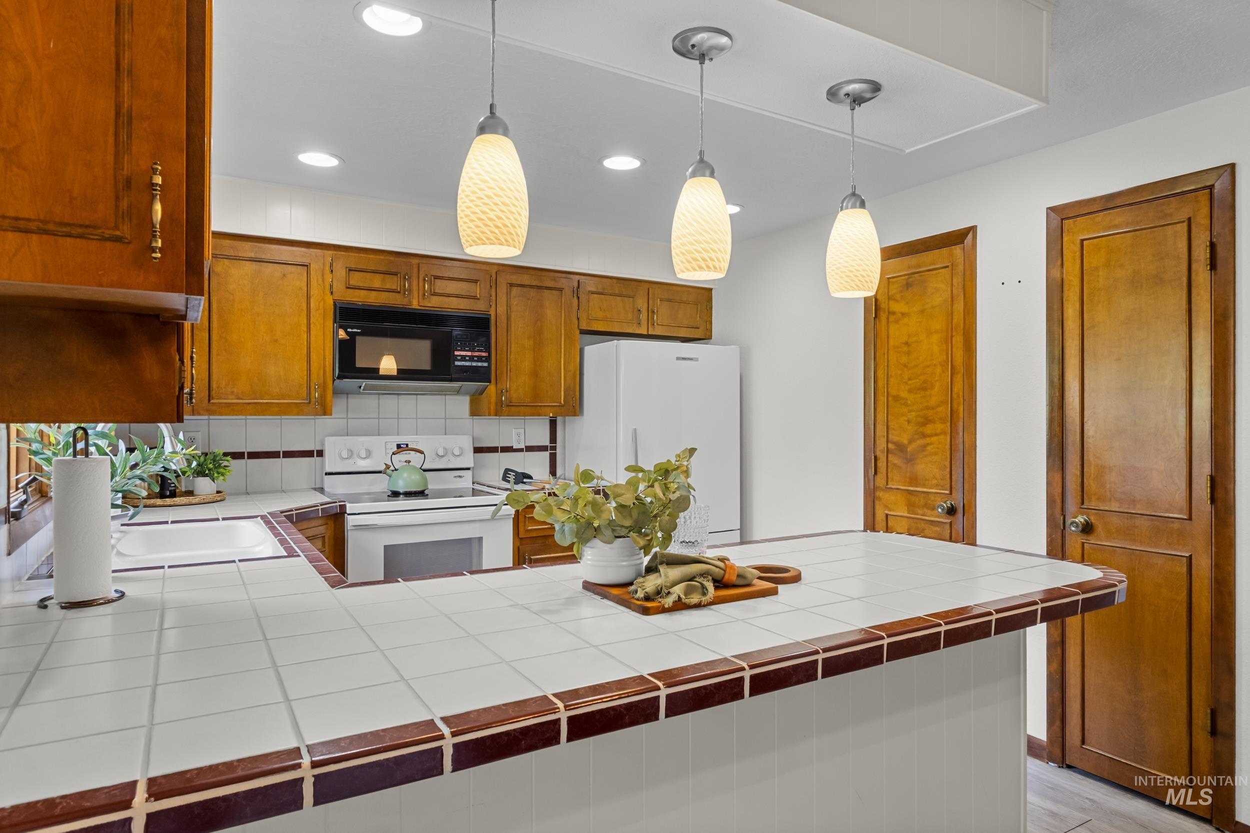 Kitchen with tile countertops, brown cabinetry, white appliances, backsplash, and recessed lighting