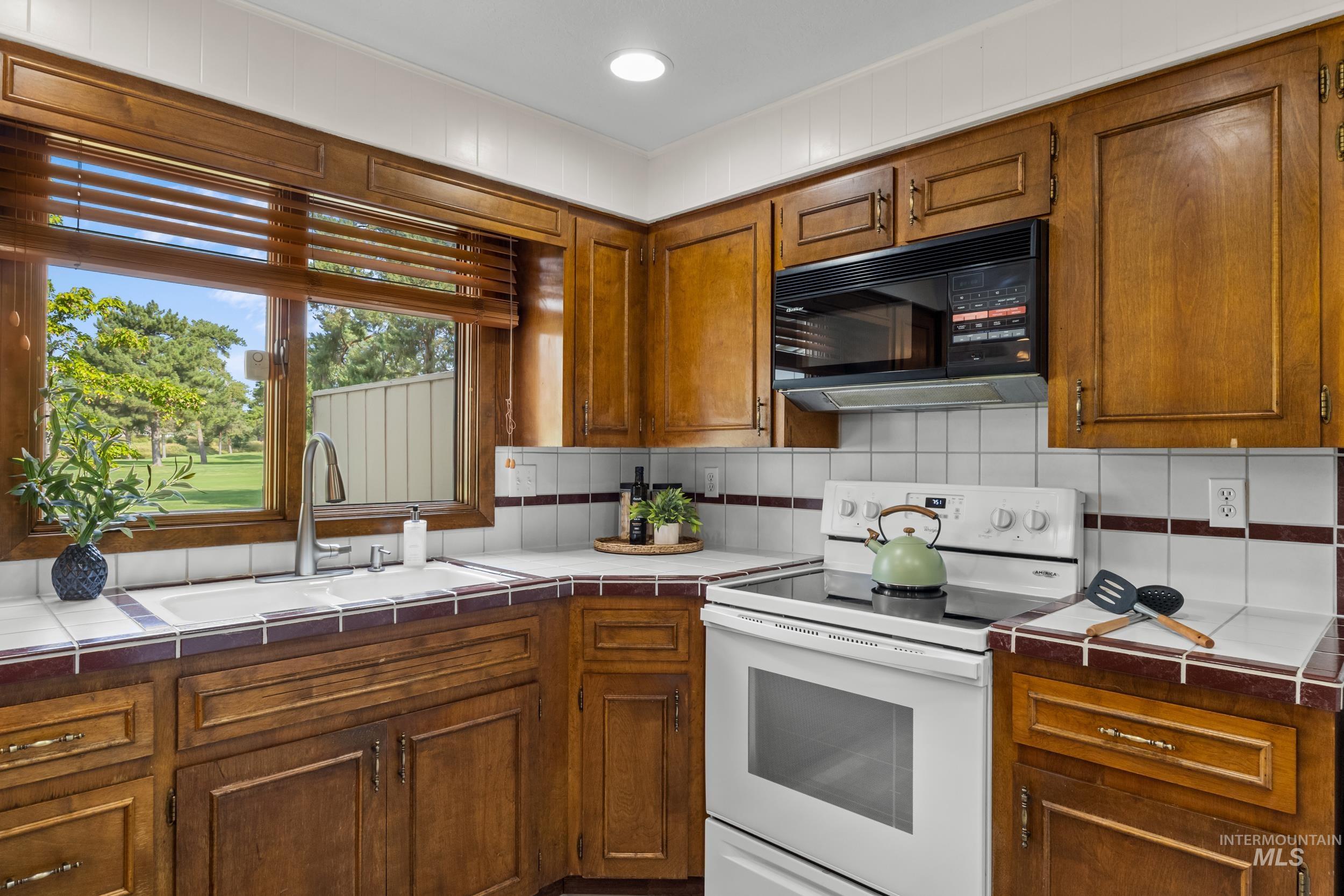 Kitchen with tile countertops, electric range, black microwave, tasteful backsplash, and brown cabinetry