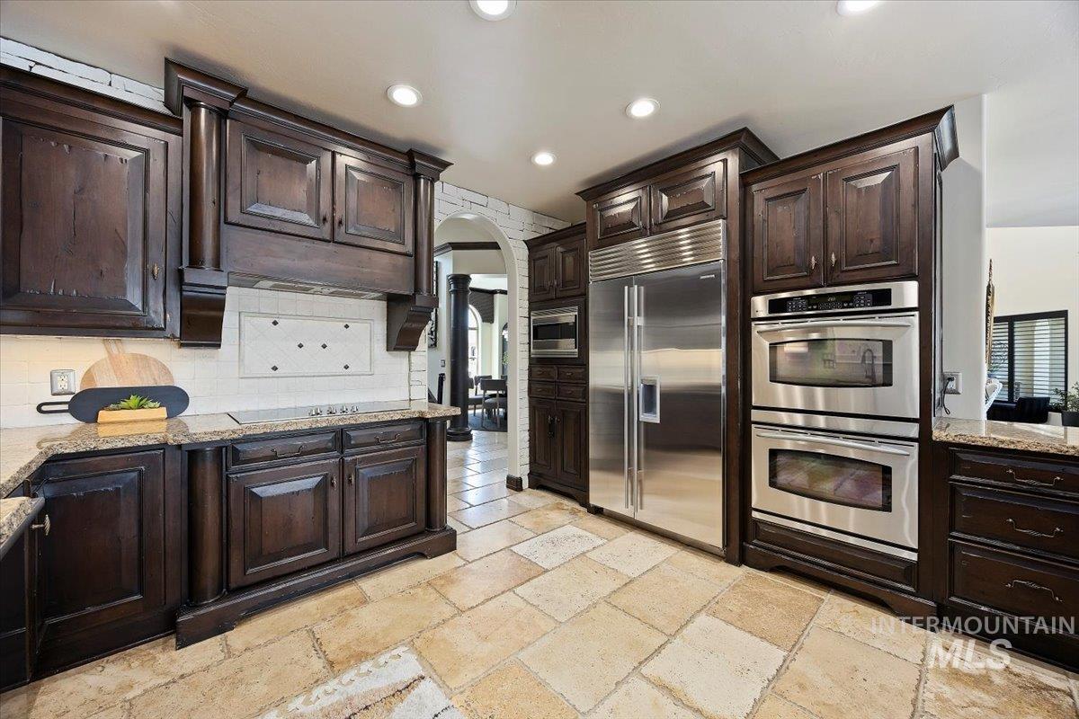 Kitchen with built in appliances, dark brown cabinetry, tasteful backsplash, light stone countertops, and stone tile flooring