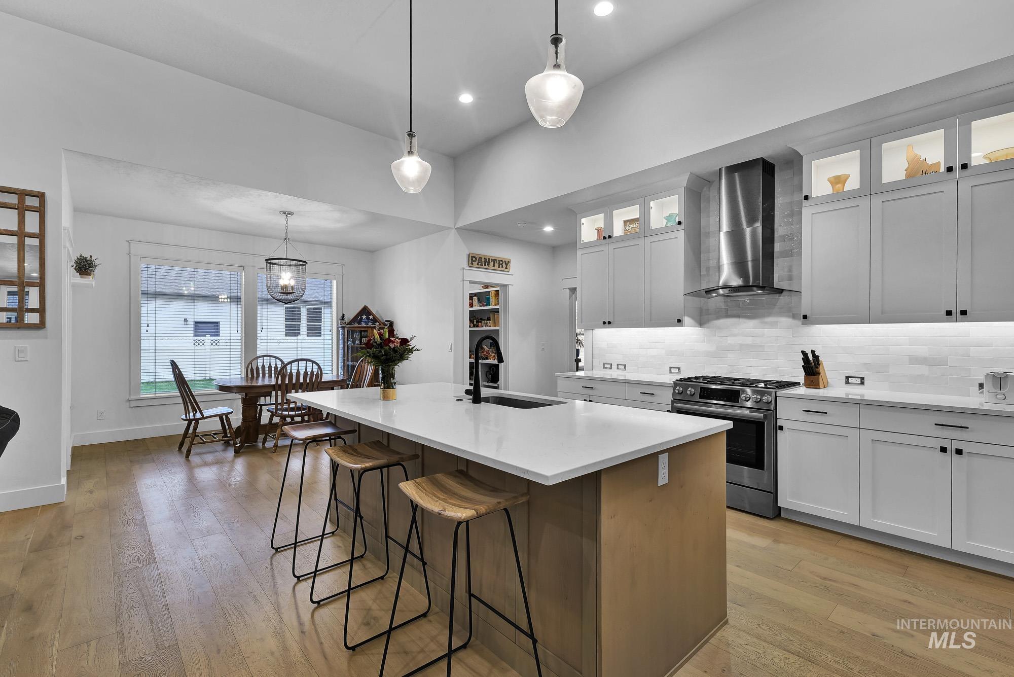 Kitchen with gas range, wall chimney range hood, a center island with sink, white cabinetry, and pendant lighting